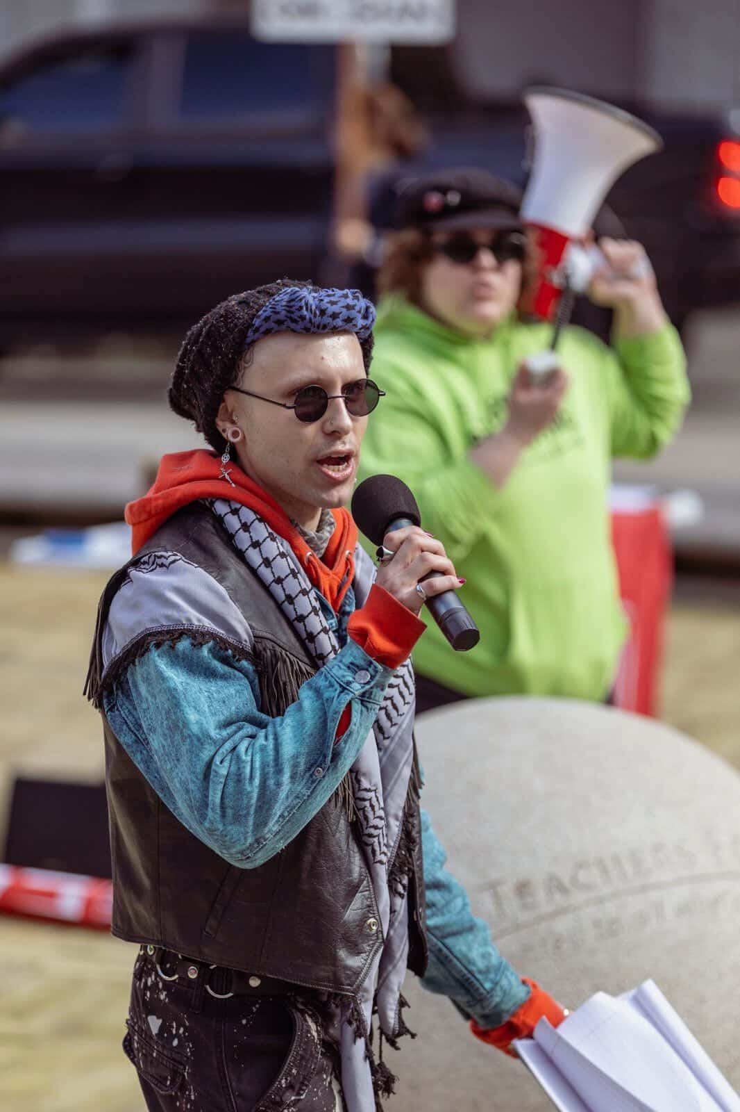 Person speaking into microphone at protest, wearing sunglasses, denim jacket, and headscarf, with another person in green holding a megaphone in background.