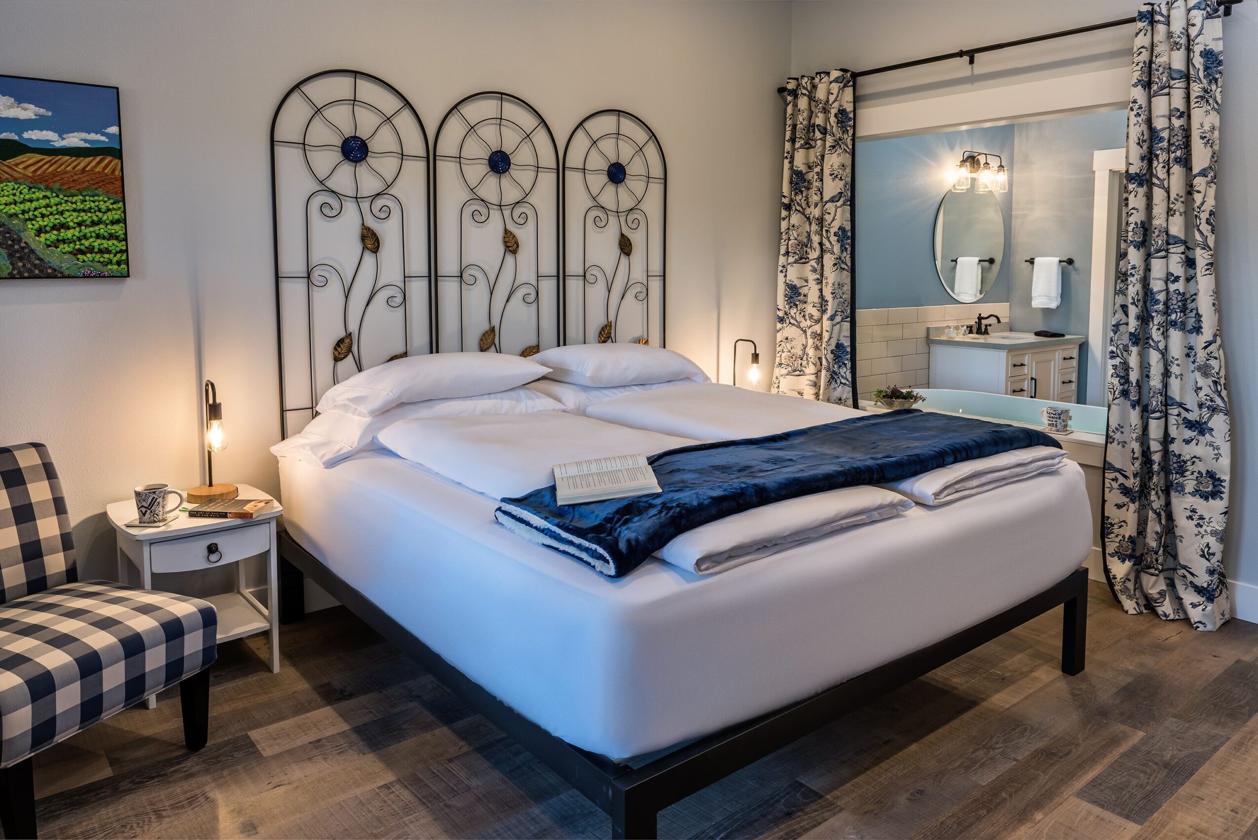 Bedroom with a large bed, decorative headboard, side table, armchair, and a mirror reflecting a dresser, curtains, and wall art.