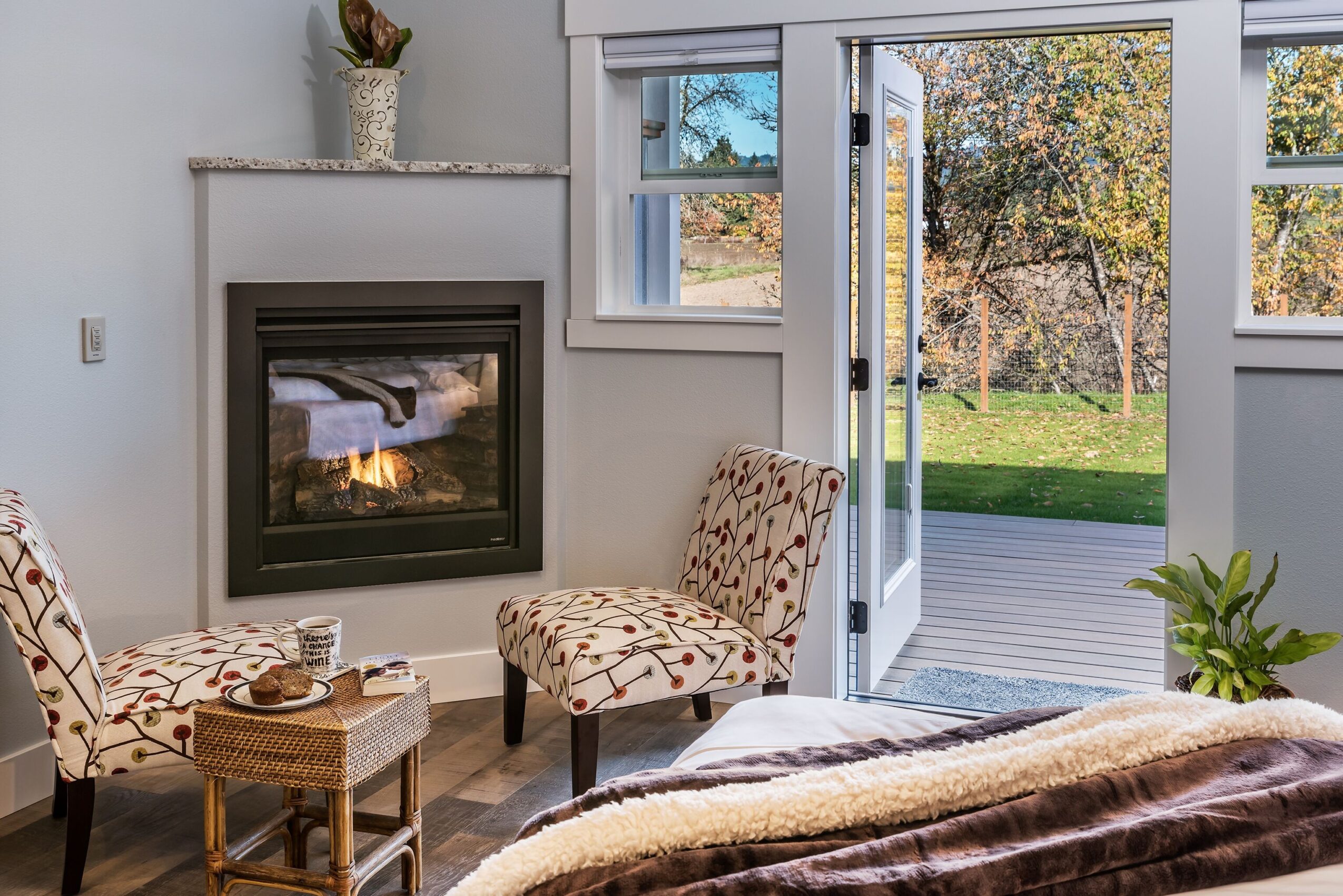 Living room with fireplace, two patterned chairs, open door to outdoor deck, and a plant near the door.