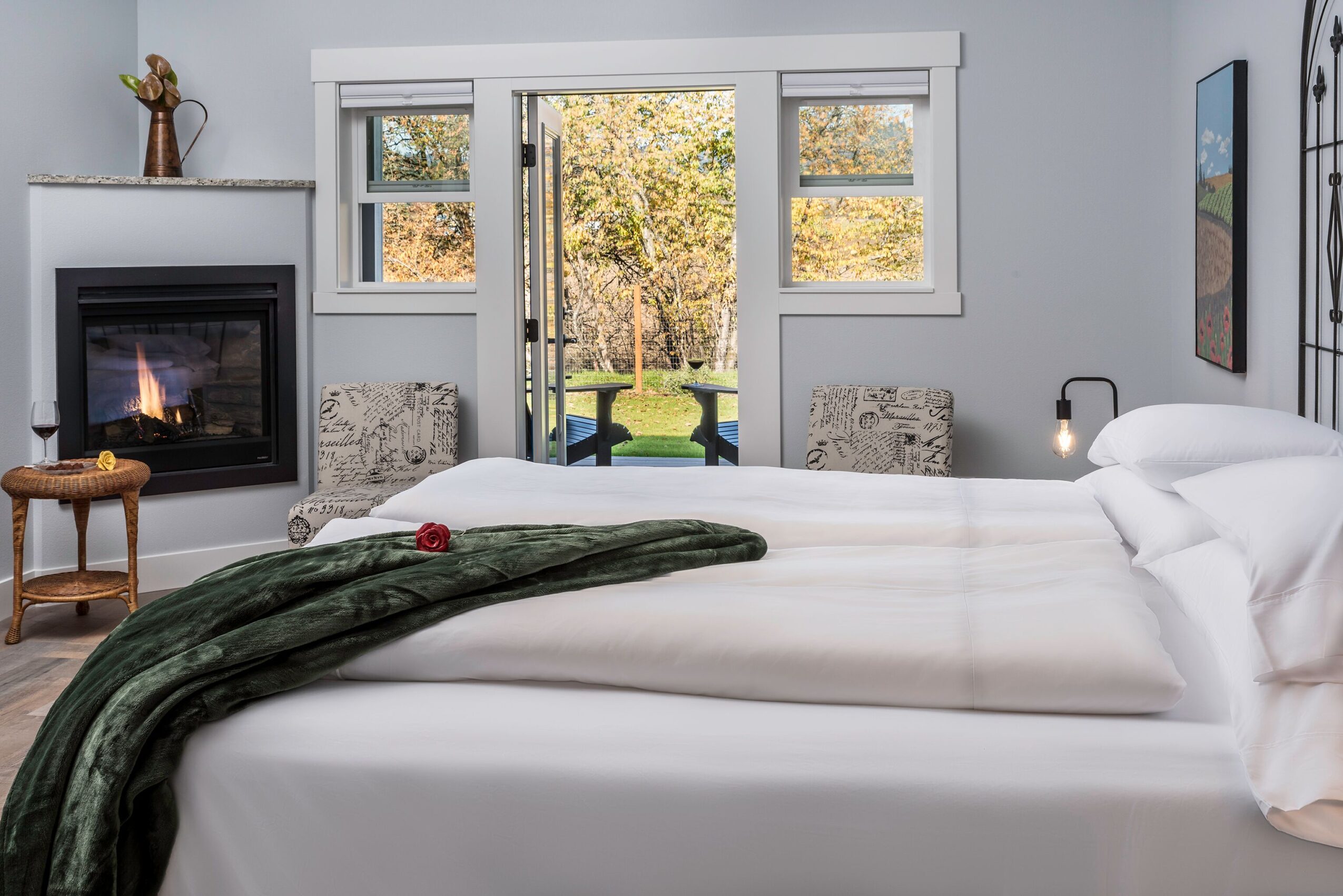 Bedroom with a bed, fireplace, and sliding glass door leading outside, with two chairs and a view of trees.