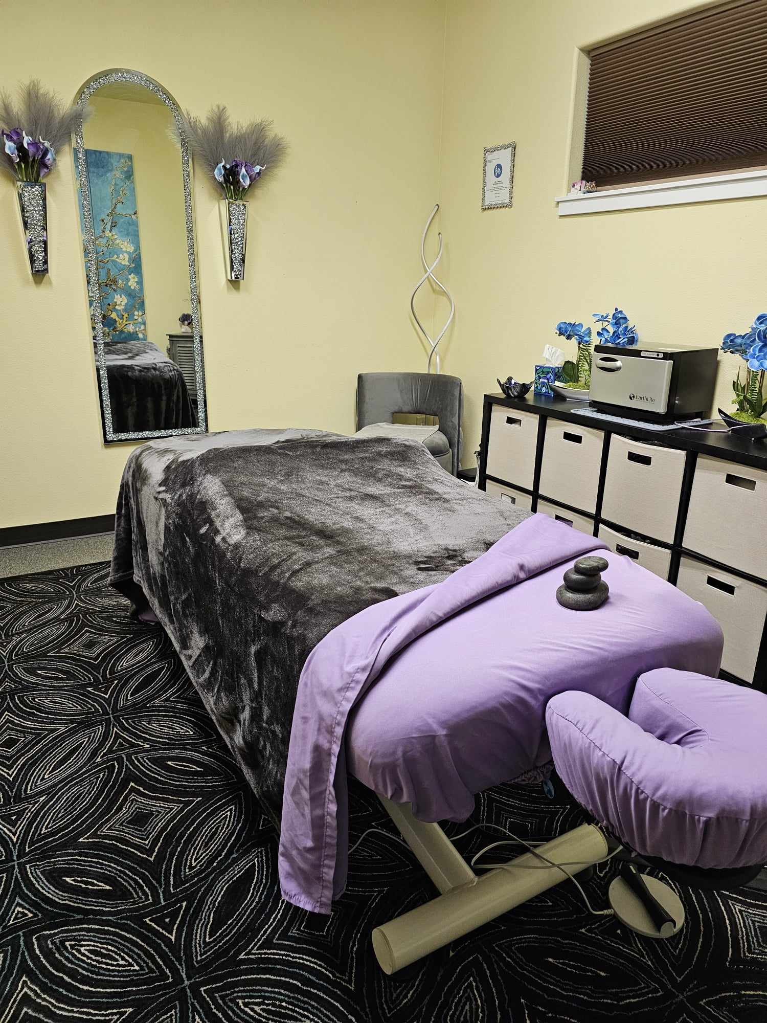 Massage table with purple sheet and remote, in a room with yellow walls, mirror, and storage cabinet, floral decorations visible.