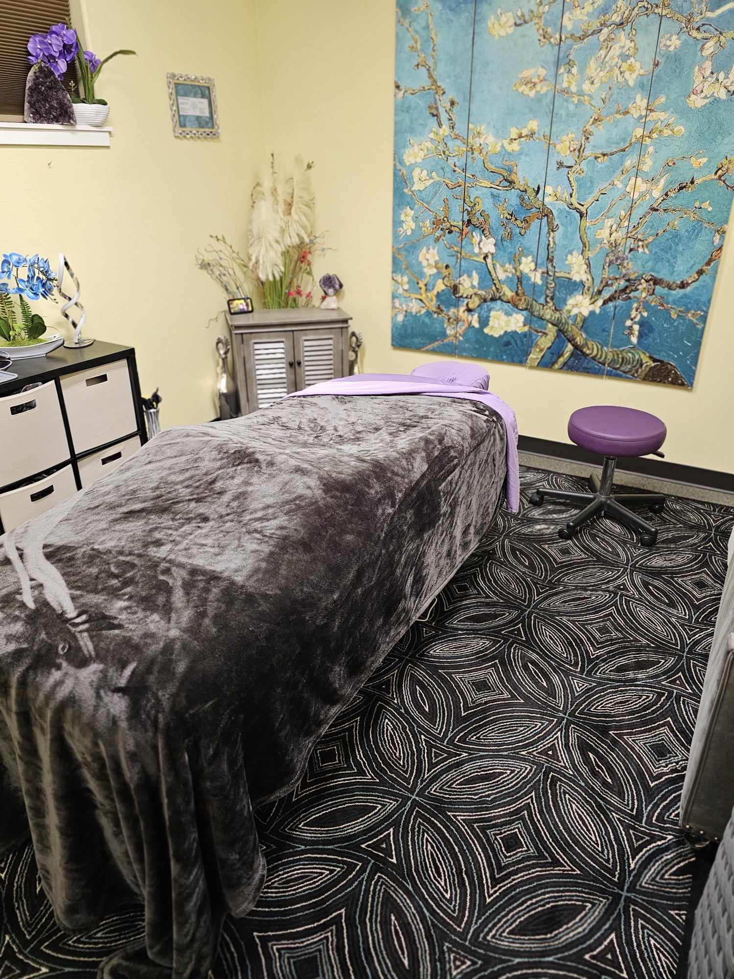 Massage table covered with gray blanket in a room with colorful wall art and patterned carpet.
