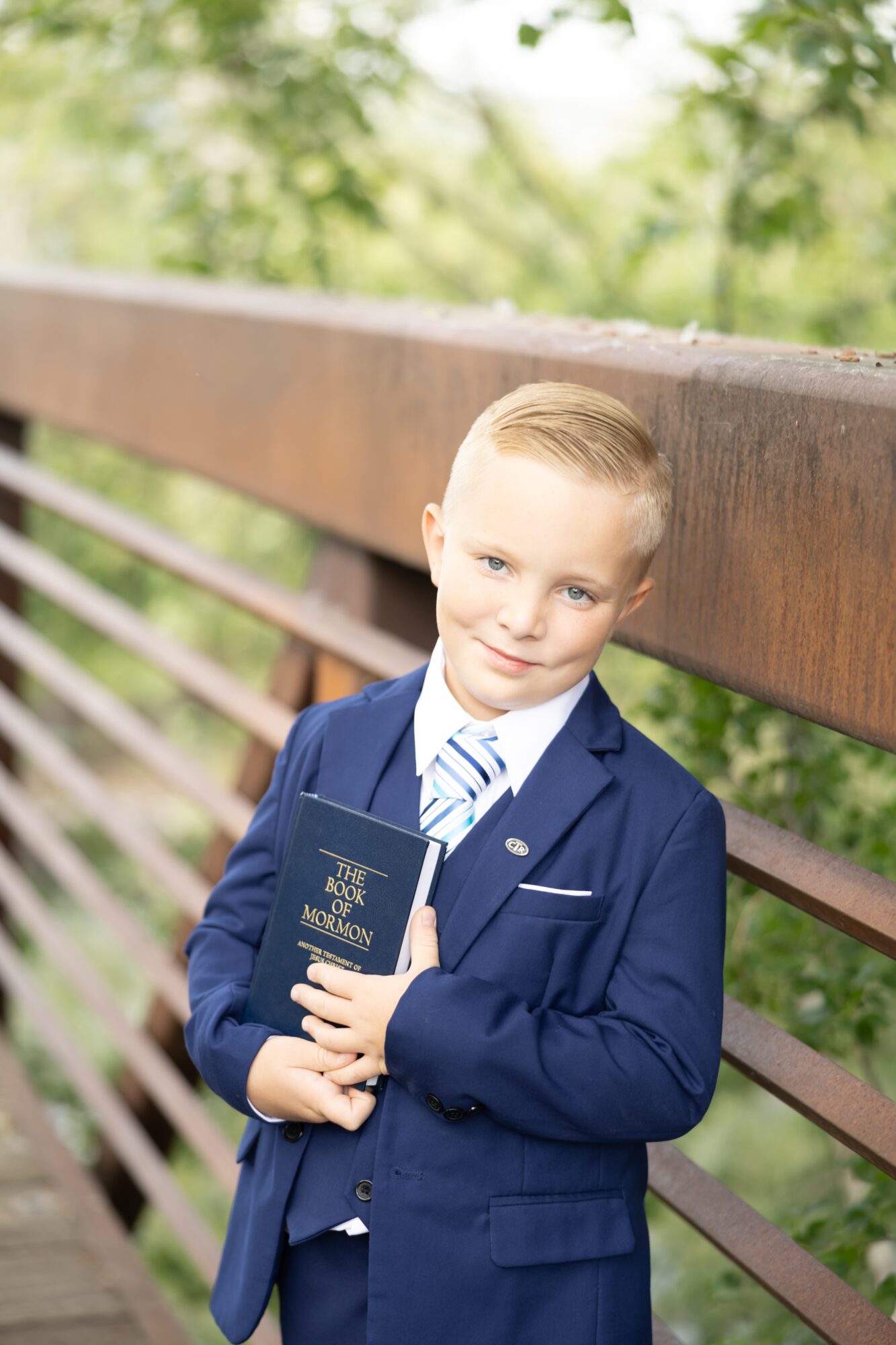 Young boy in a suit holding a diploma, standing outdoors near a wooden fence with trees in the background.