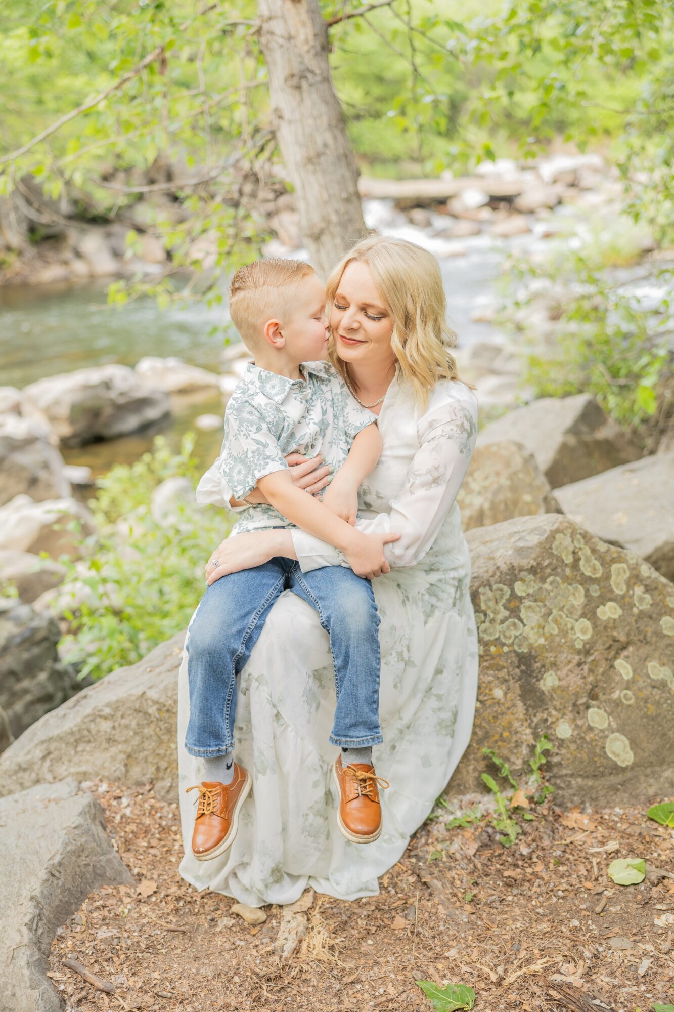 Woman holding a child on a large rock outdoors with trees and a stream in background.