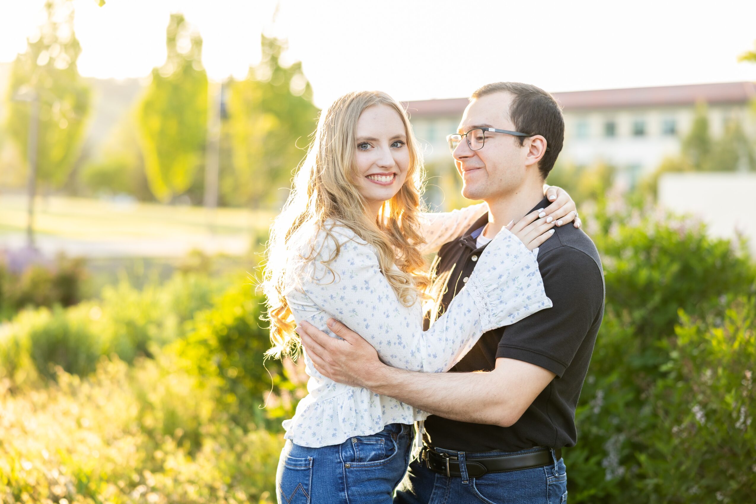 A smiling couple embracing outdoors with greenery and a building in the background.