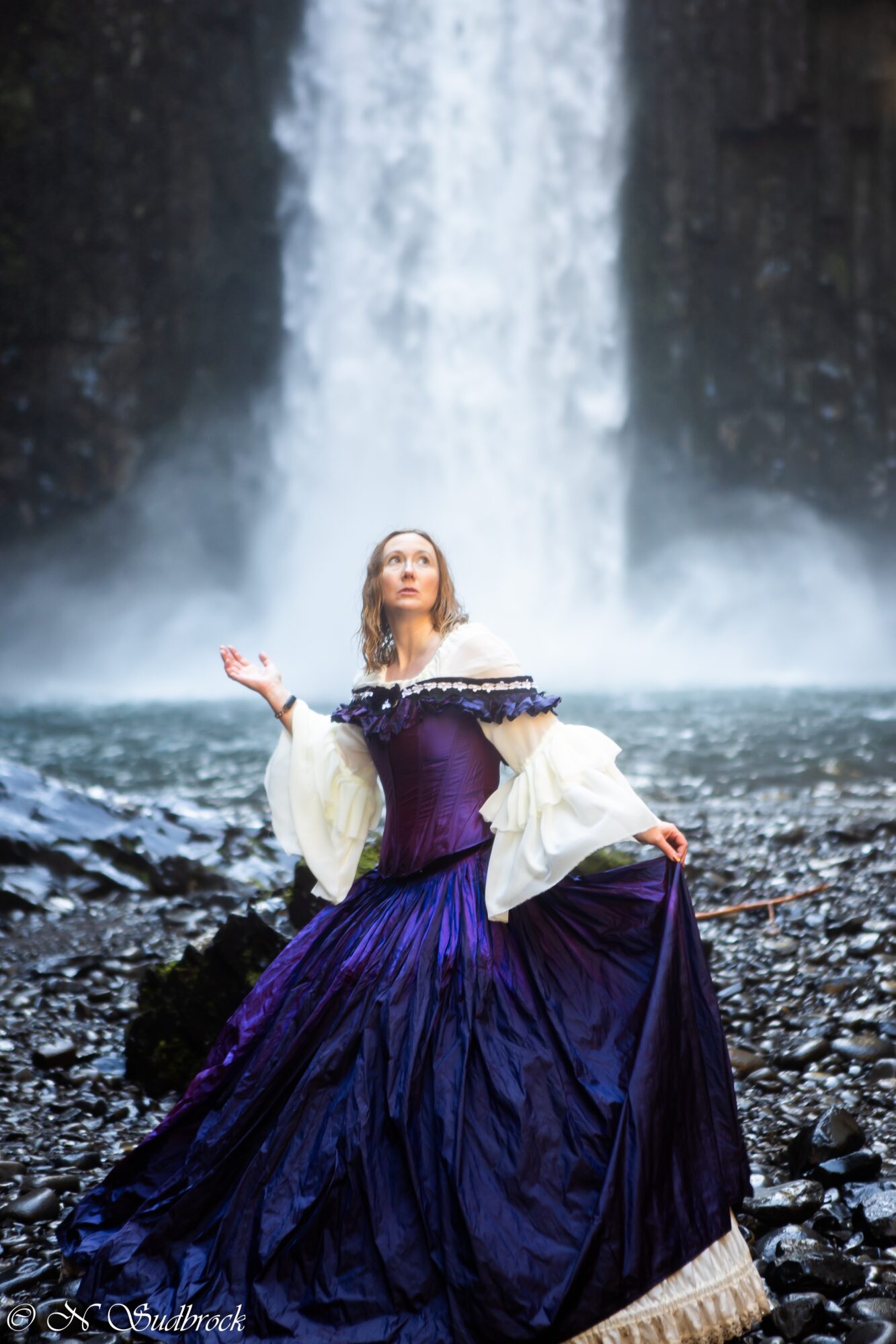 Woman in a purple gown and white shawl stands in front of a waterfall, looking upward with arms outstretched.