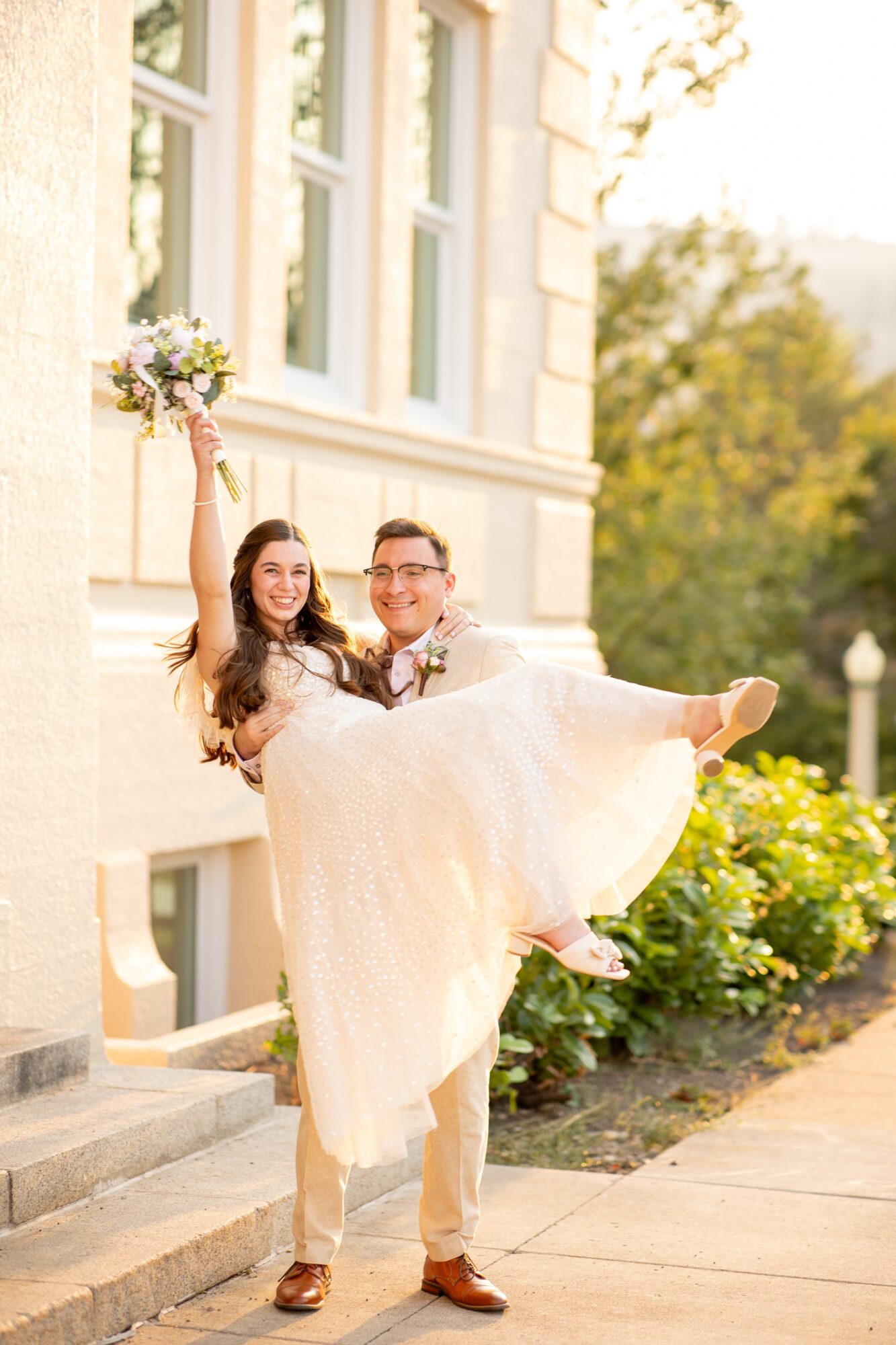 Man carrying woman in wedding dress holding bouquet outside building, sunlight, greenery, and flowers nearby.
