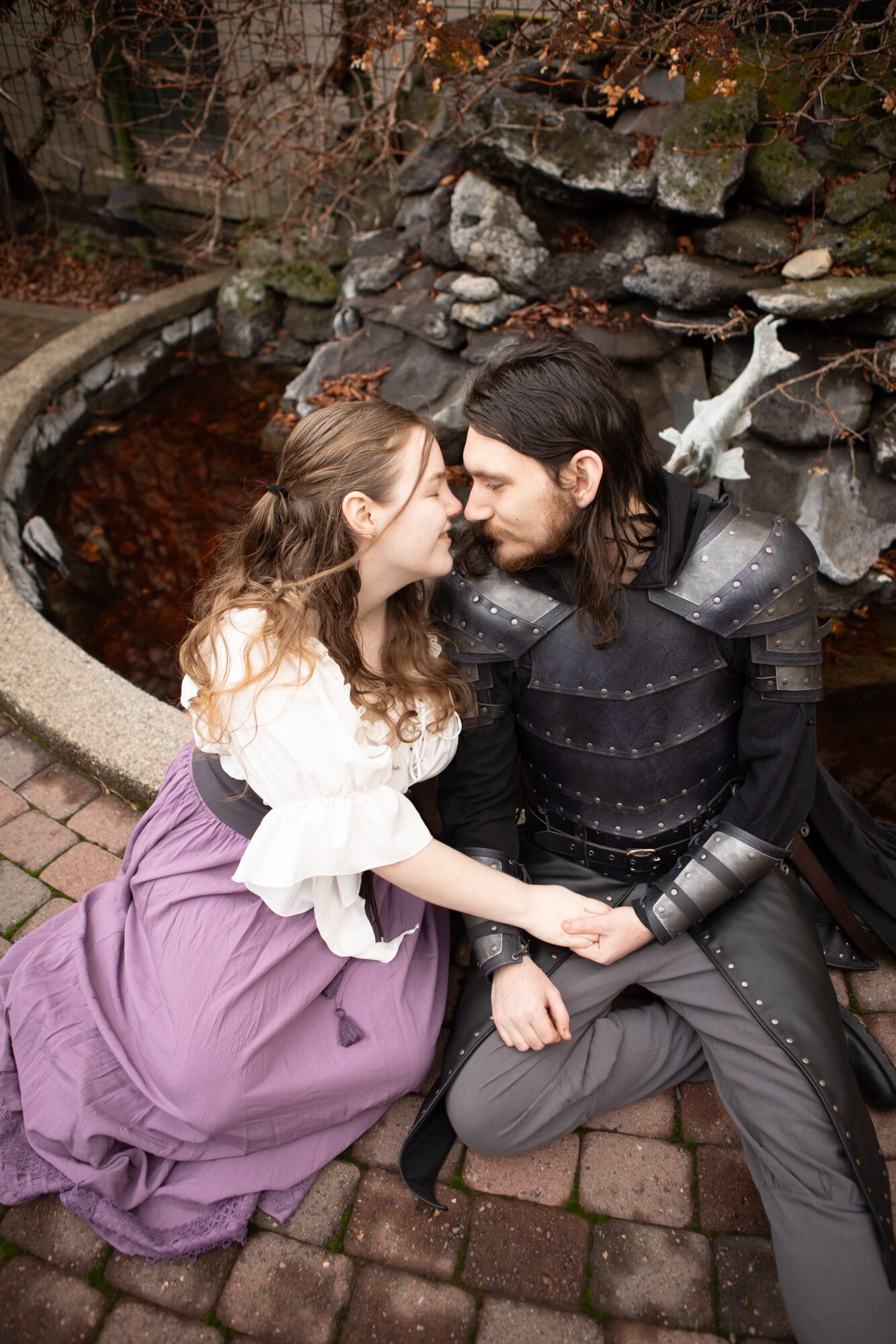 Two people in medieval costumes sit close, holding hands, near a stone fountain with a dragon sculpture in the background.
