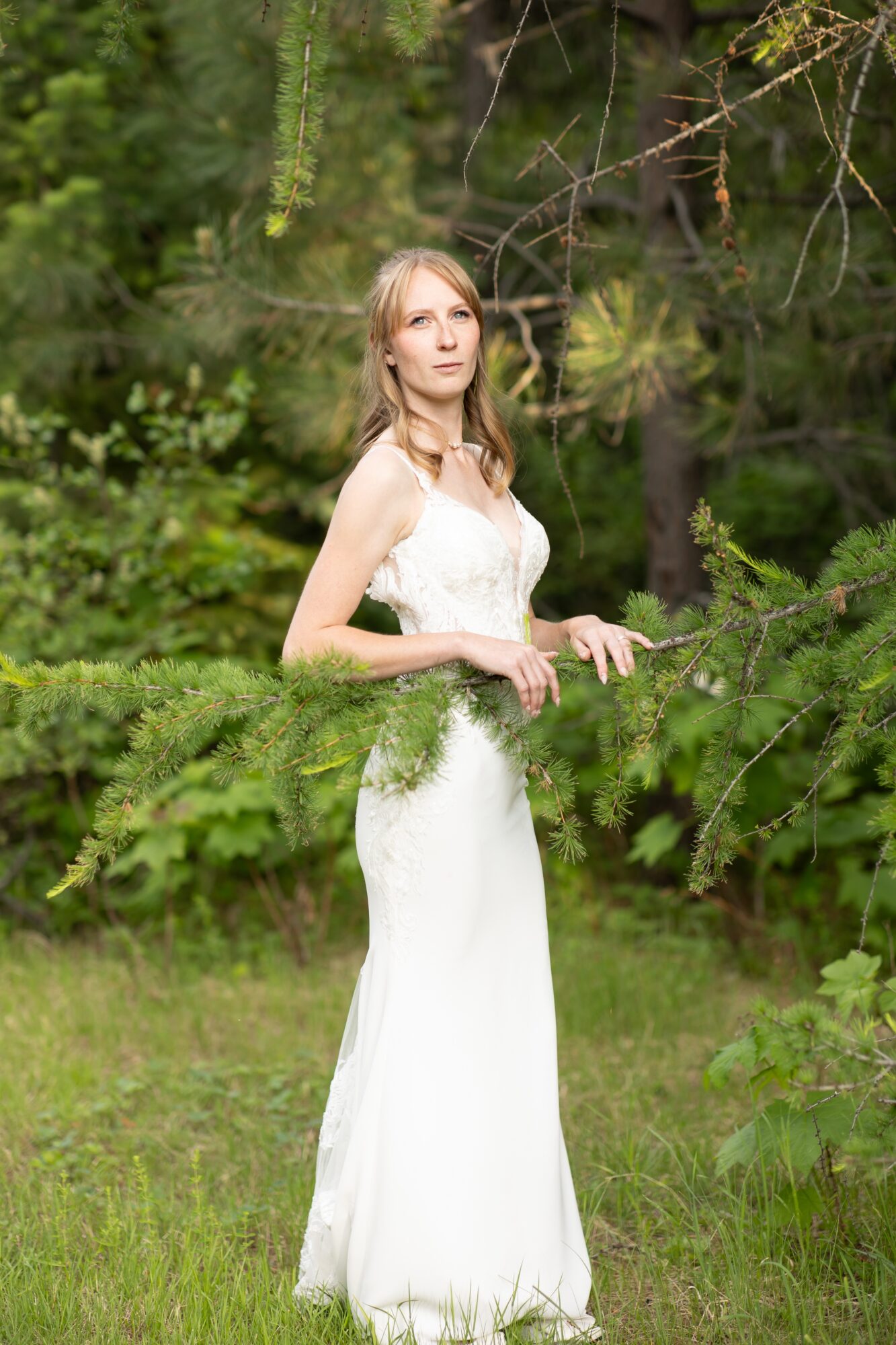 Woman in a white dress standing outdoors among green trees and bushes, looking into the distance.
