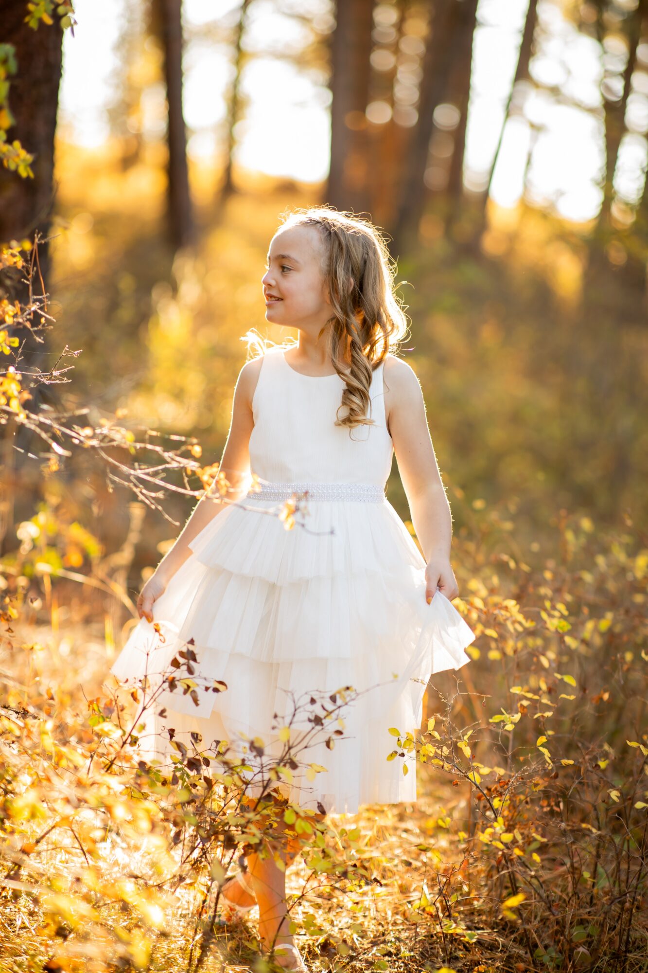 Young girl in a white dress standing outdoors in a forest with sunlight filtering through trees.