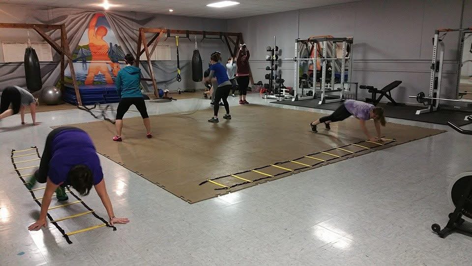 People exercising in a gym with equipment and a wooden floor, some in plank positions, others doing different stretches.