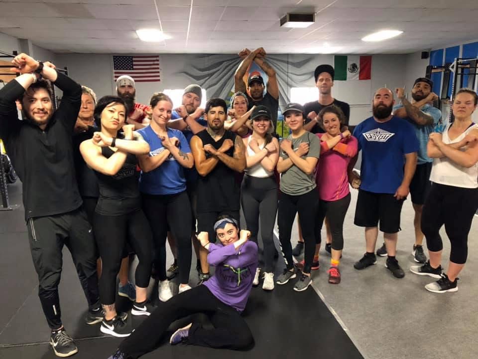 Group of people in a gym, some crossing arms, posing for a photo, with flags and gym equipment in background.
