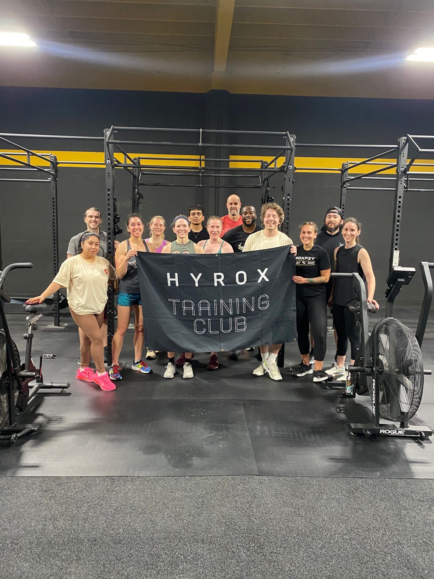 Group of people in gym, some holding weights, others sitting or standing, with gym equipment in background.