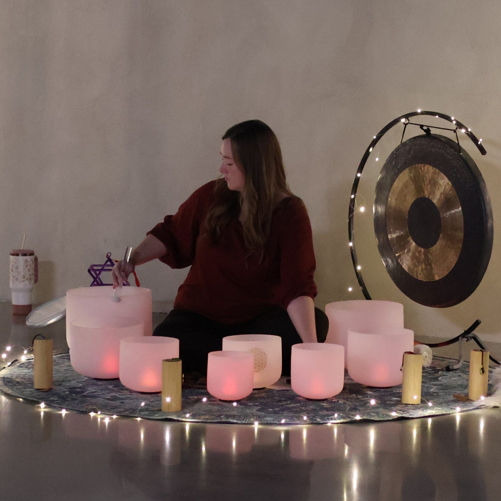 Woman sitting among illuminated singing bowls and candles, with a large gong on the wall behind her.
