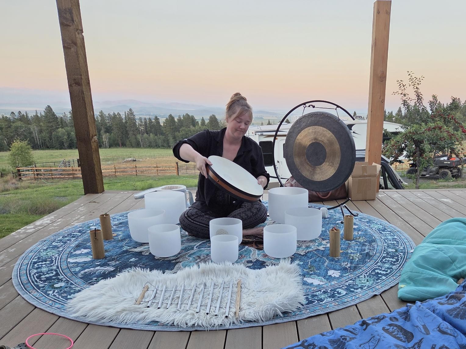 Person sitting on a round rug outdoors, playing a gong with a mallet, surrounded by cups and candles, with trees and sky in background.