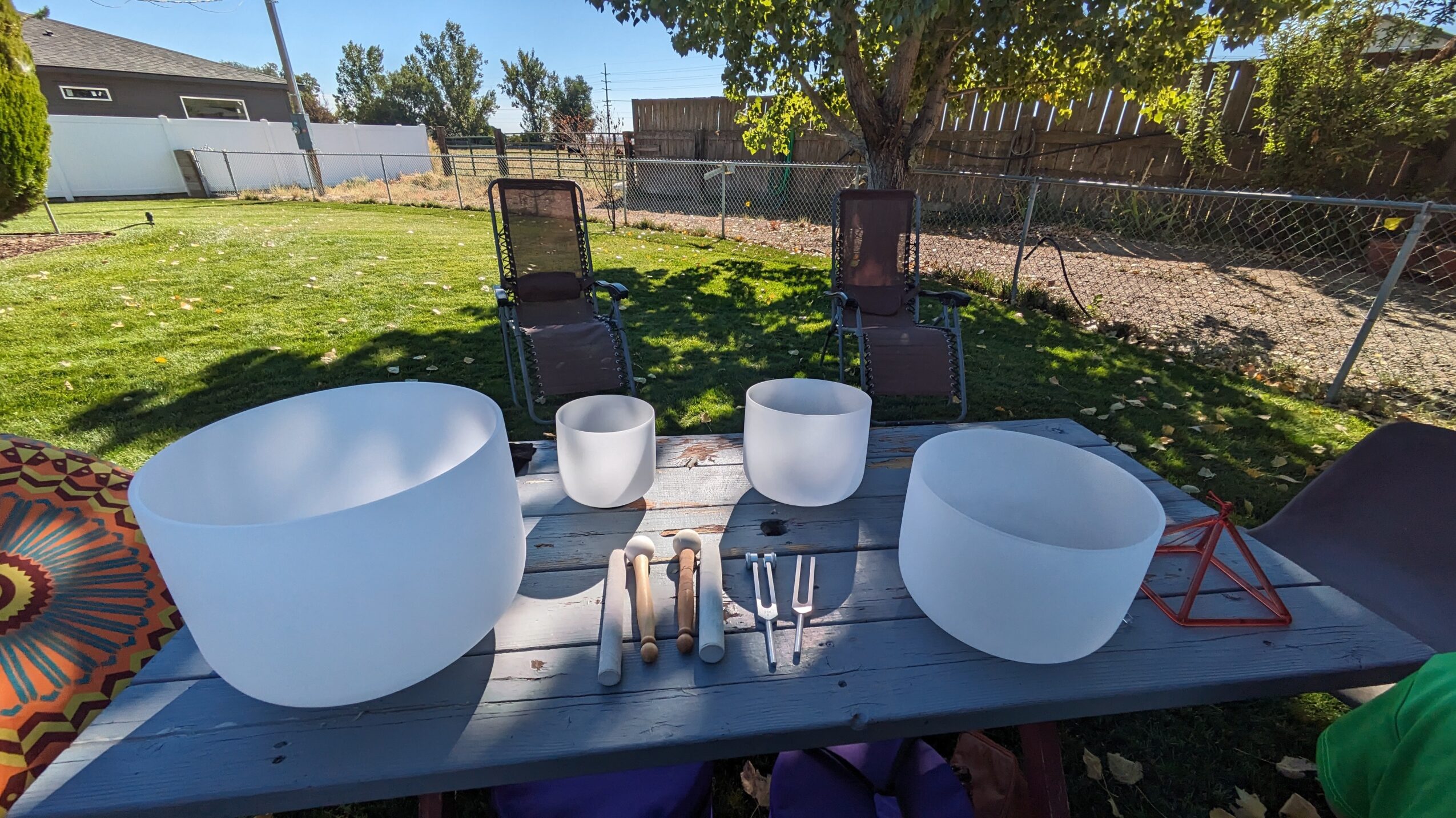 Outdoor scene with two large white singing bowls, two smaller bowls, and various percussion instruments on a table, with chairs and trees in background.