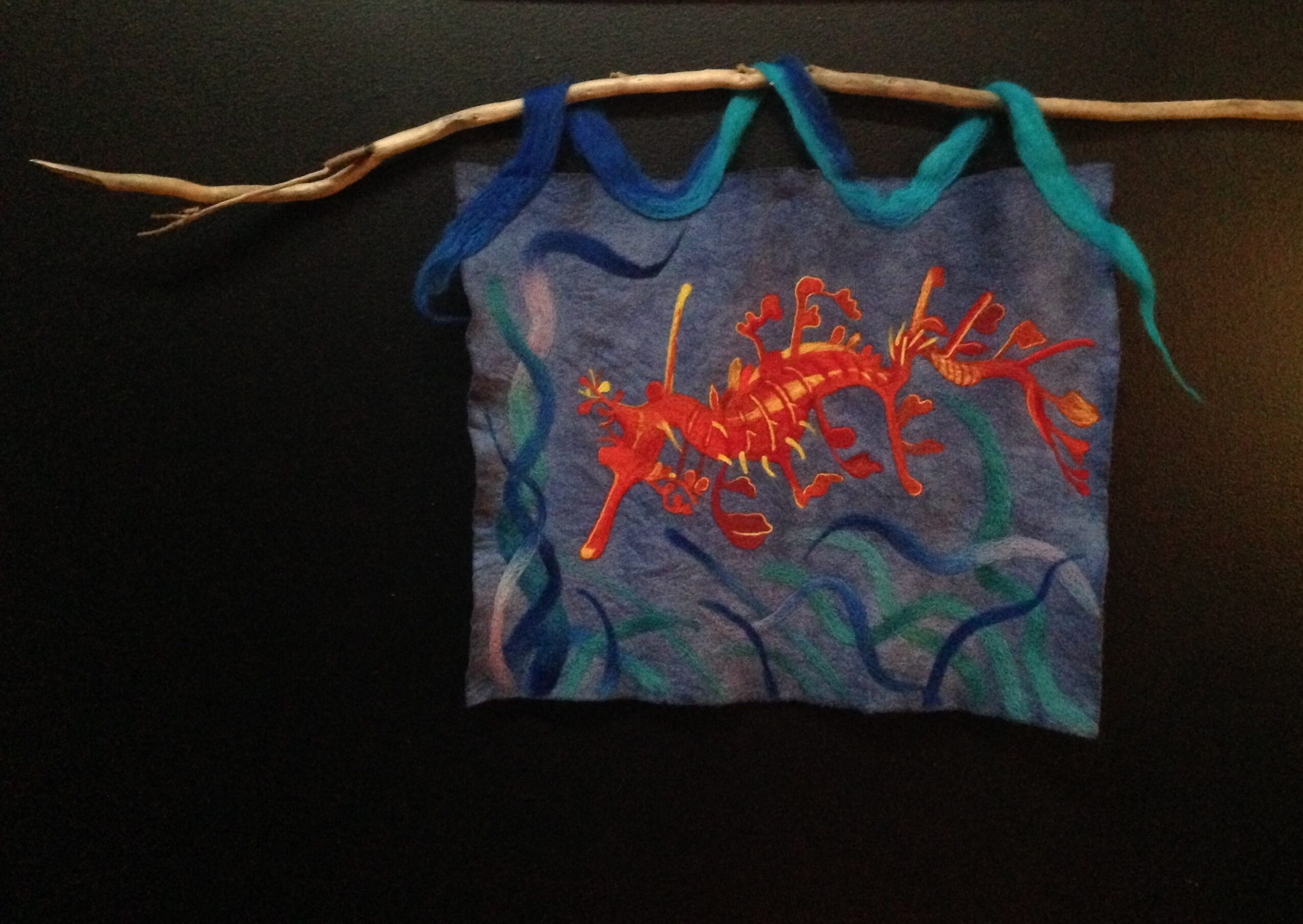 Fabric with a red and orange embroidered creature, hanging from a branch with blue and green fabric strips, against a dark background.