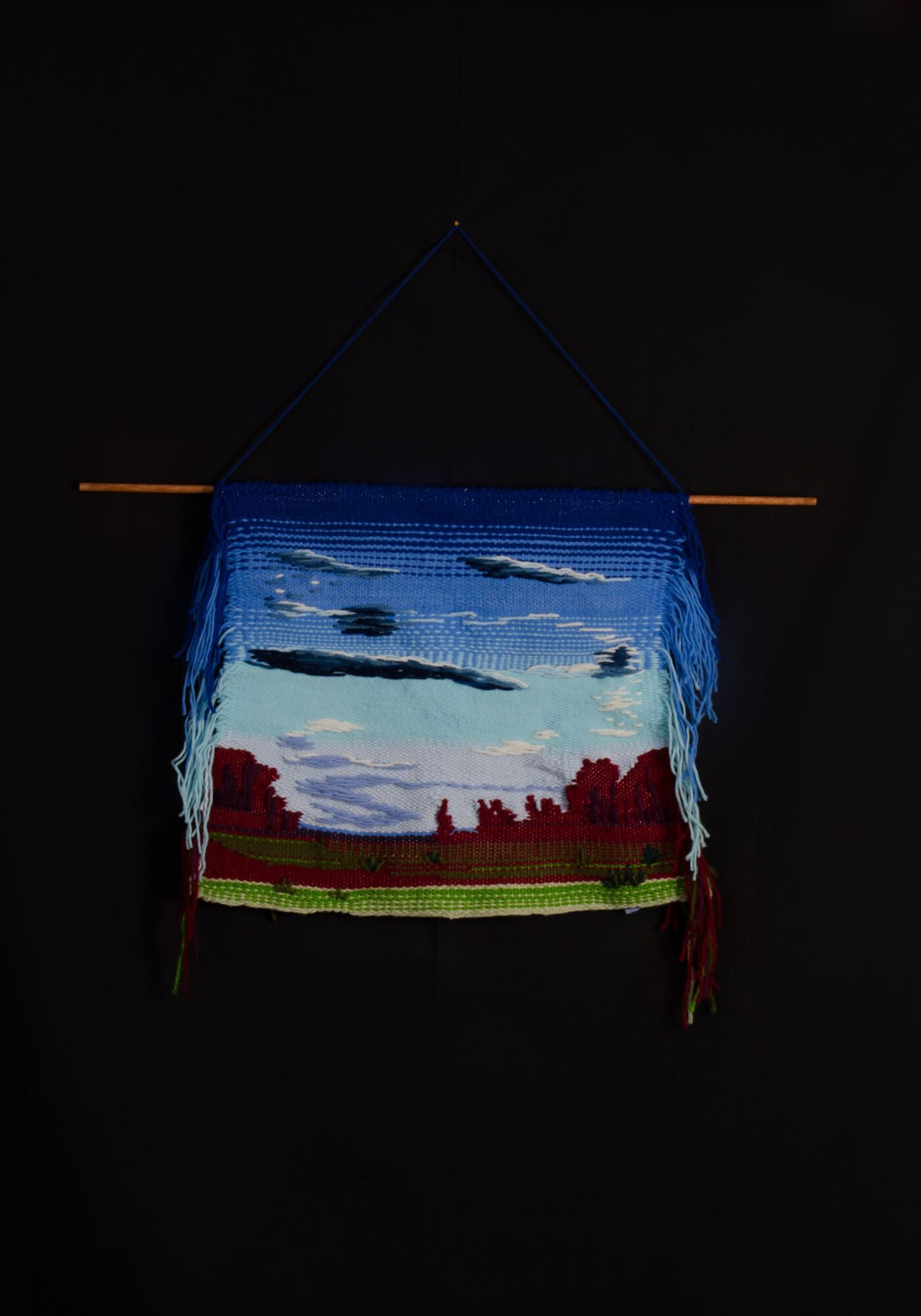 Colorful woven wall hanging with landscape scene, hanging on a black background, with a horizontal wooden rod and a string.