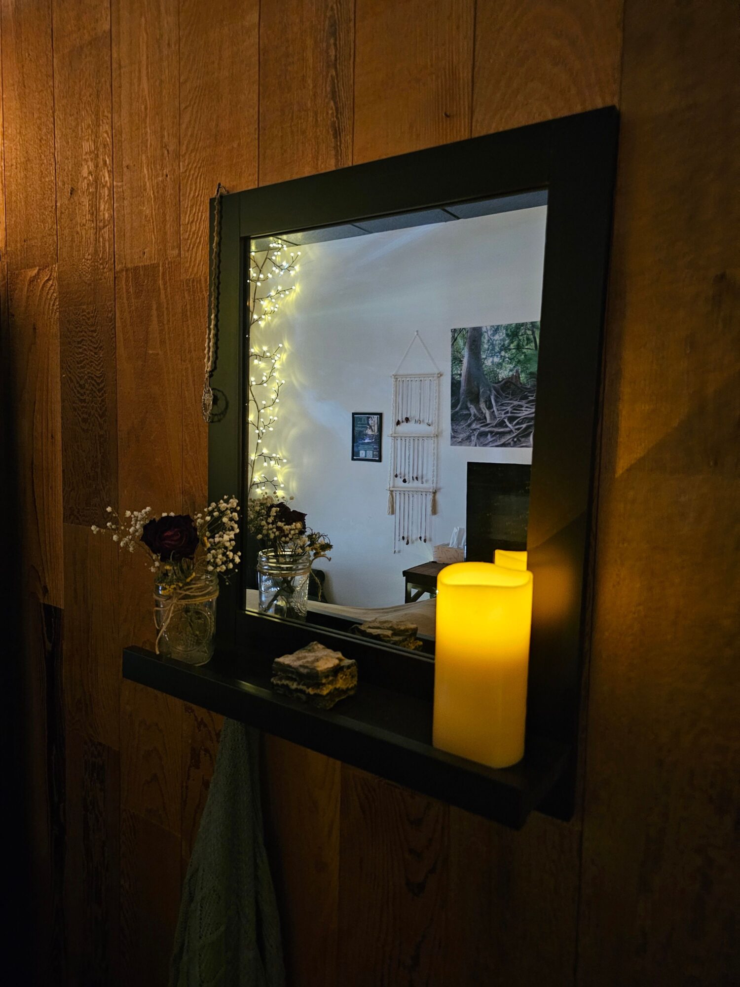 A framed picture leaning against a wooden wall with a yellow lamp, flowers, and decorative items on a black table.