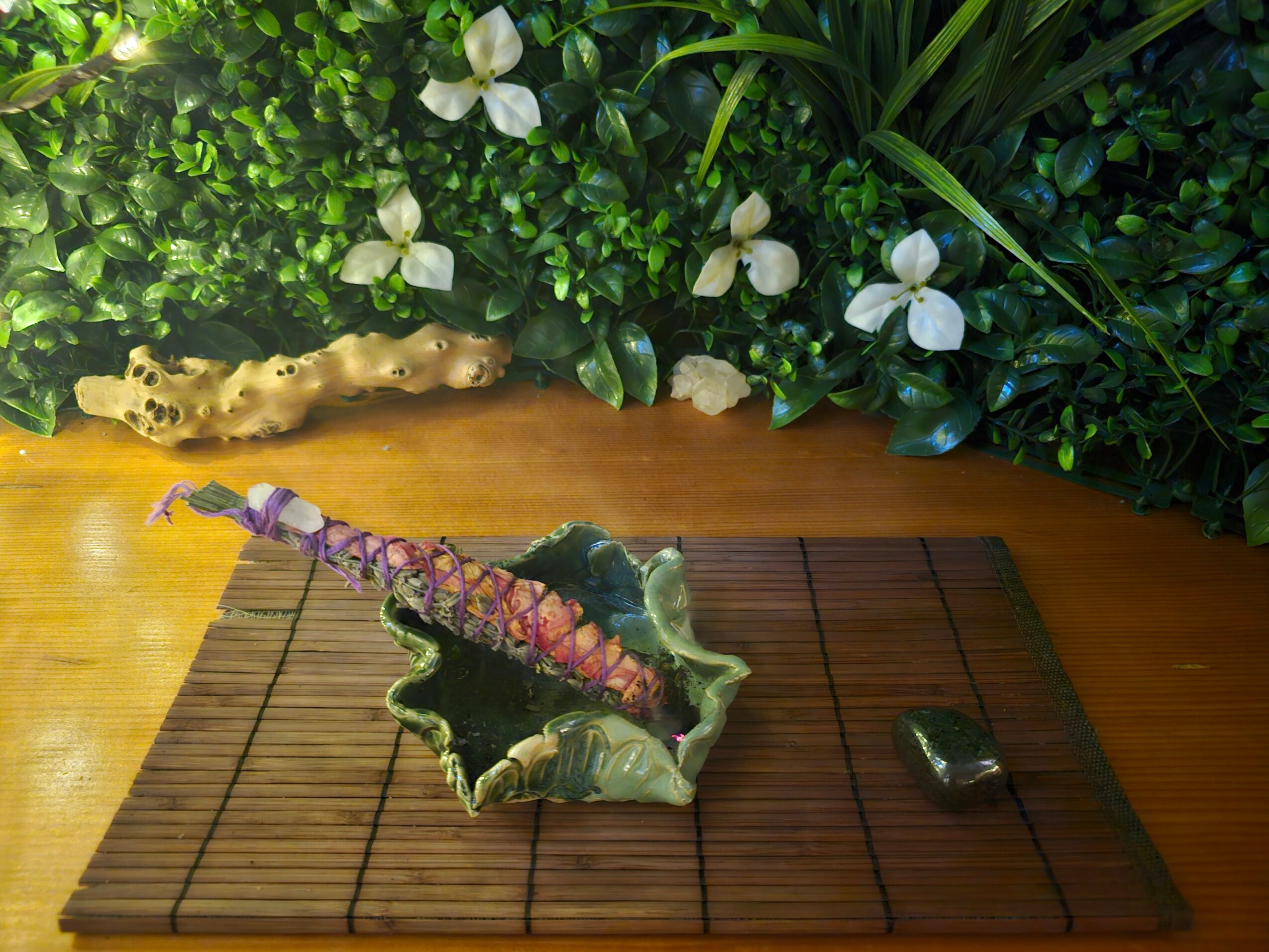 A plant with a purple and pink flower on a decorative green and beige ceramic stand on a wooden surface, with green foliage and white flowers in the background.