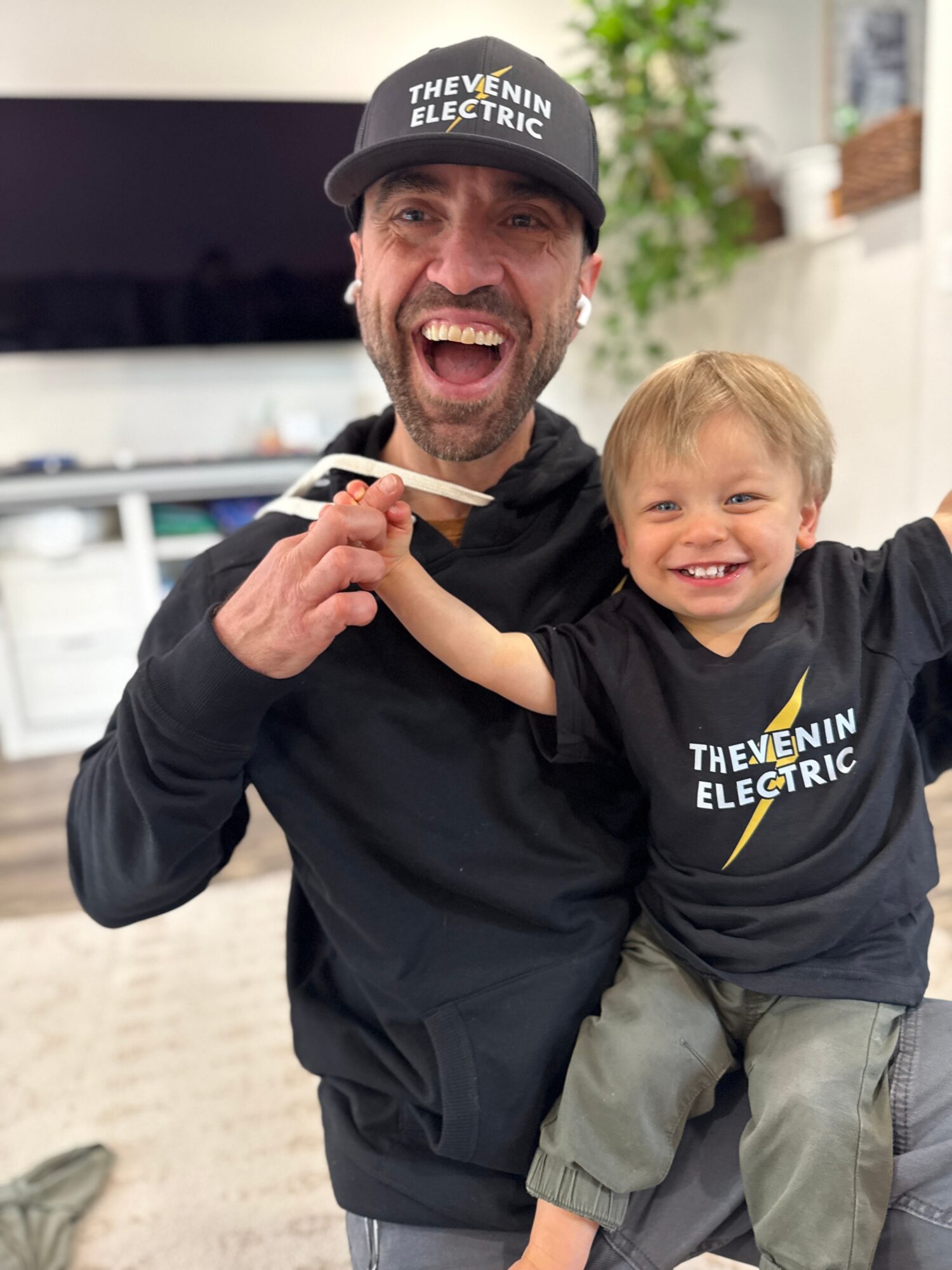 Man and child smiling, man holding a pen, both wearing black shirts and hats with 'The Venn Electric' logo, indoors.