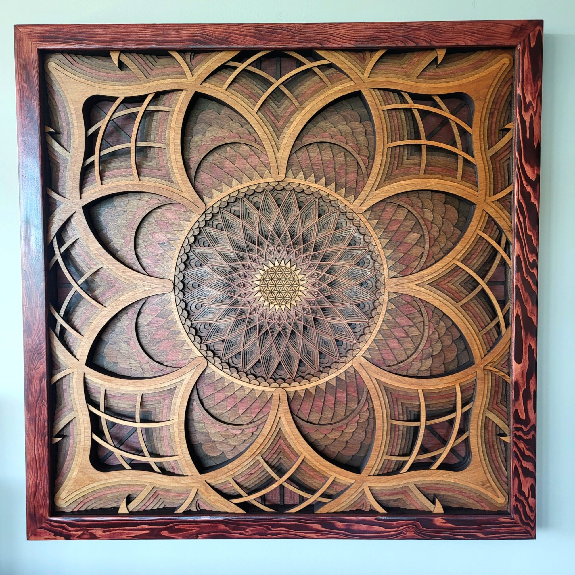 Intricate wooden geometric pattern with a central circular design, framed by a reddish-brown border, displayed on a wall.