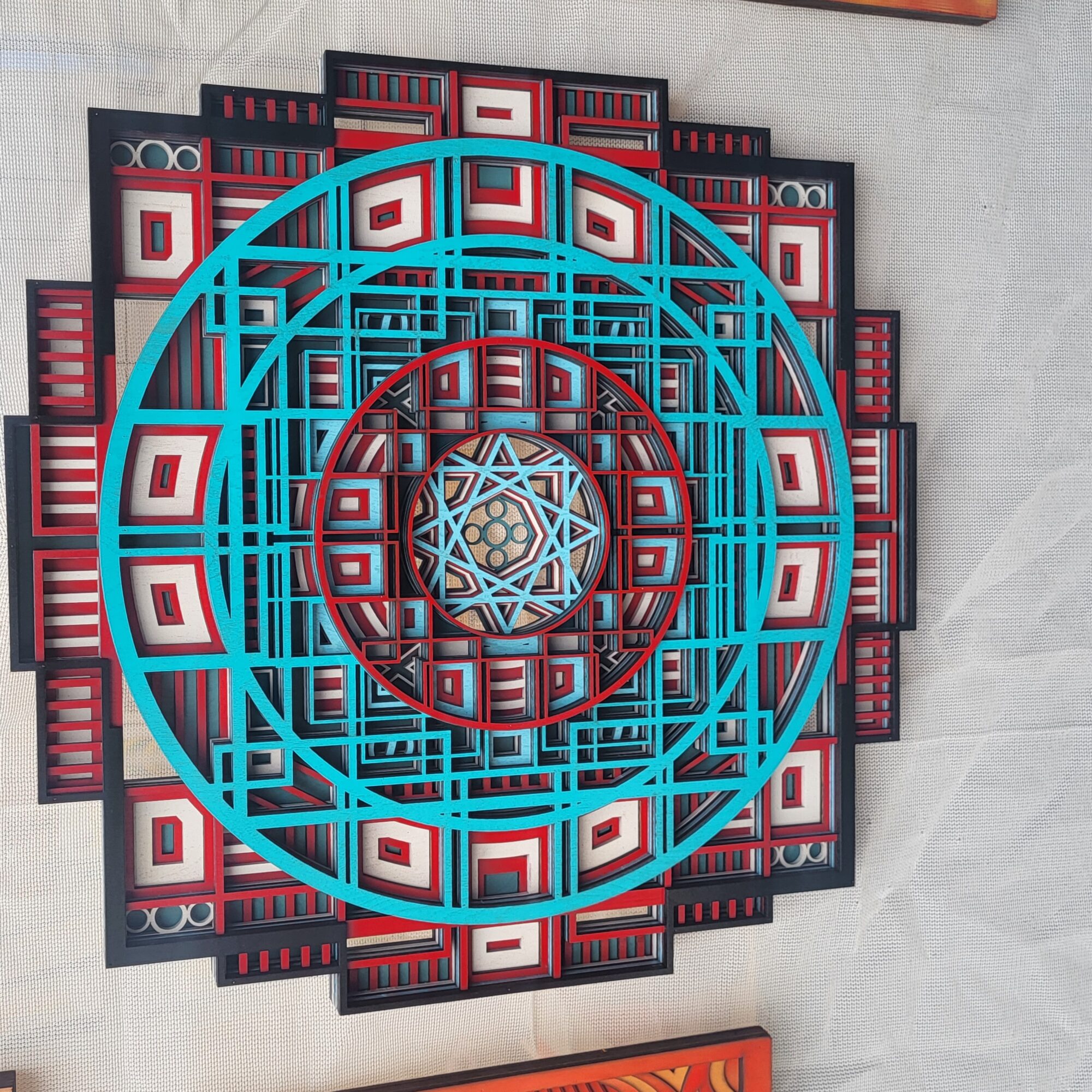 Colorful geometric wall art with concentric circles and intricate patterns in blue, red, and black, mounted on a light gray wall.
