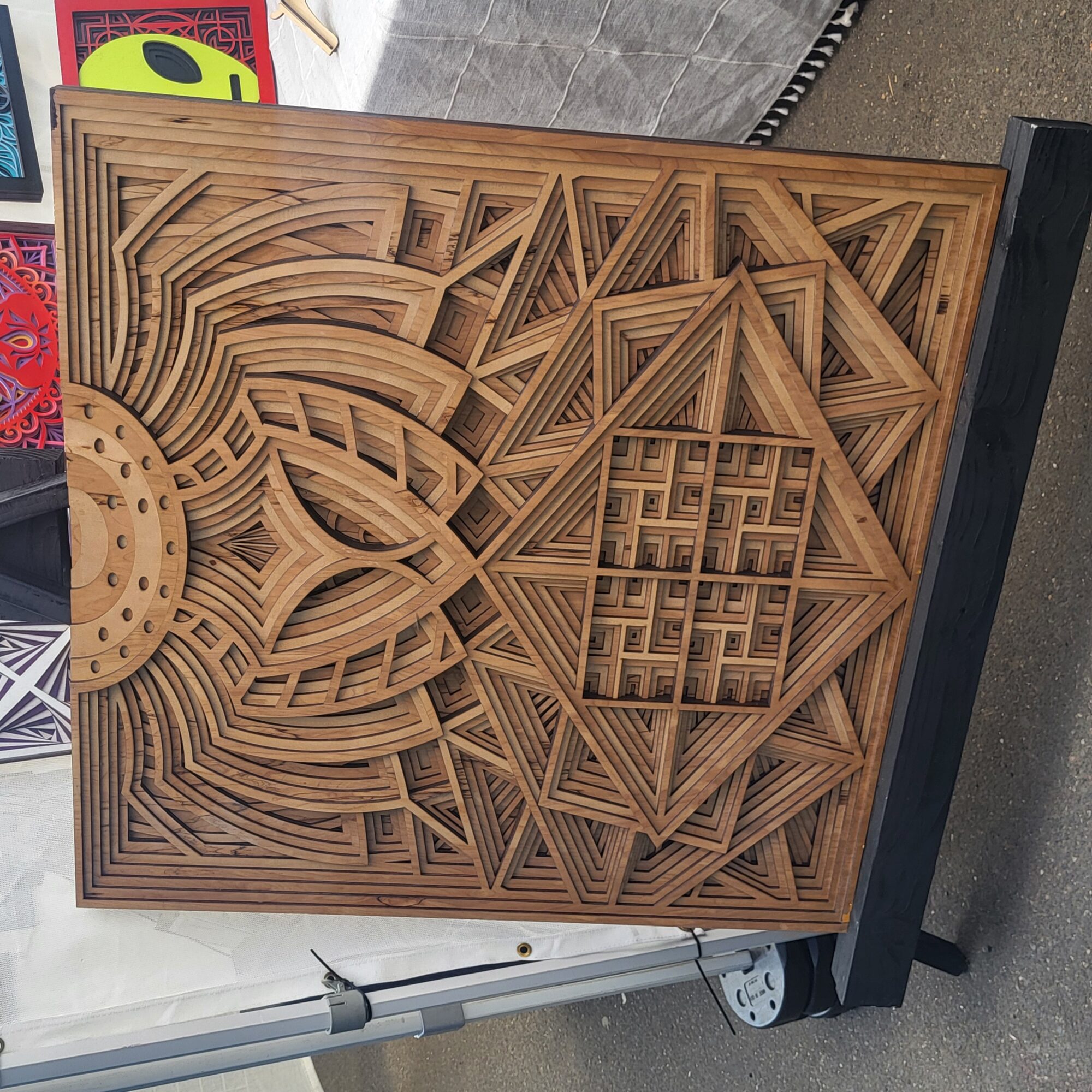 Intricate wooden geometric pattern with layered lines and shapes on a square panel, mounted on a black stand.