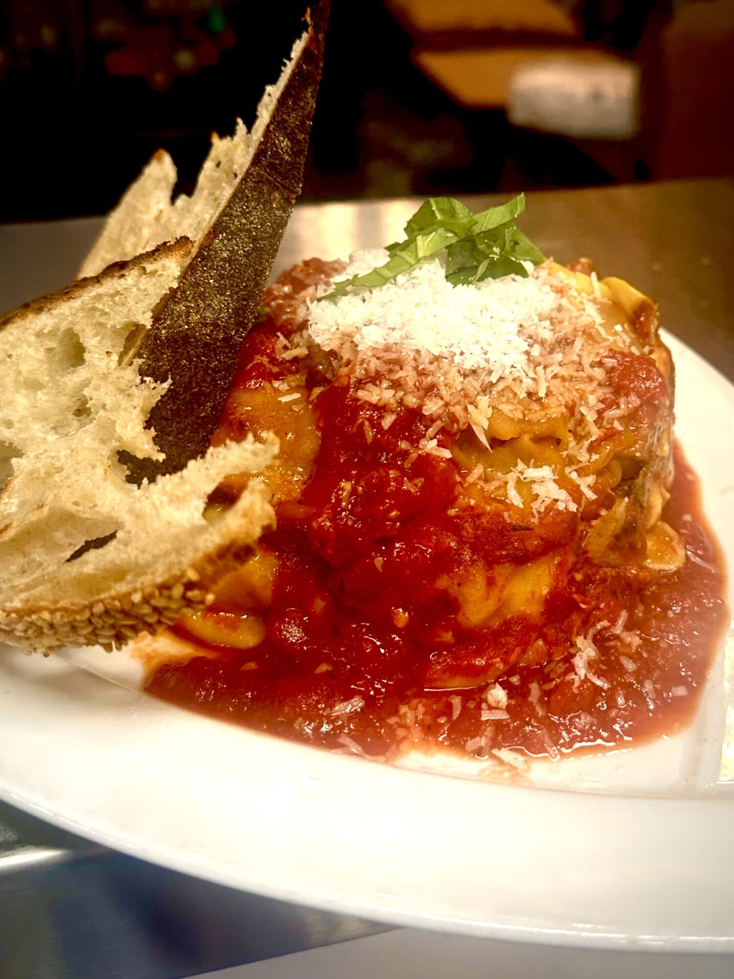 Plate with a serving of lasagna topped with cheese and herbs, with a piece of bread on the side, on a white plate.