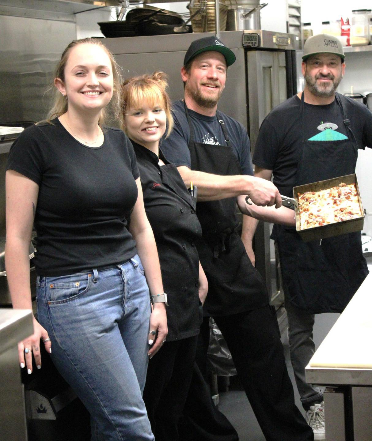 Four people in a kitchen, smiling, one holding a pizza box, casual clothing, industrial kitchen background.
