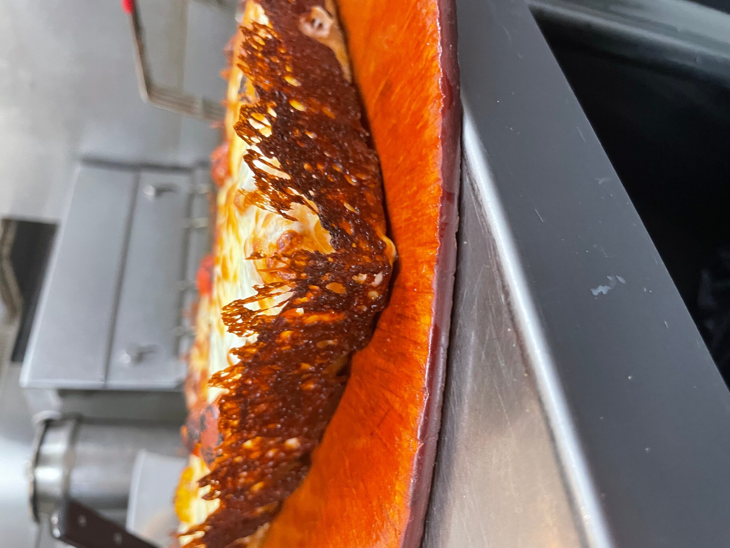 Close-up of a pizza with cheese, tomato, and a crispy topping, viewed from the side on a metal surface.
