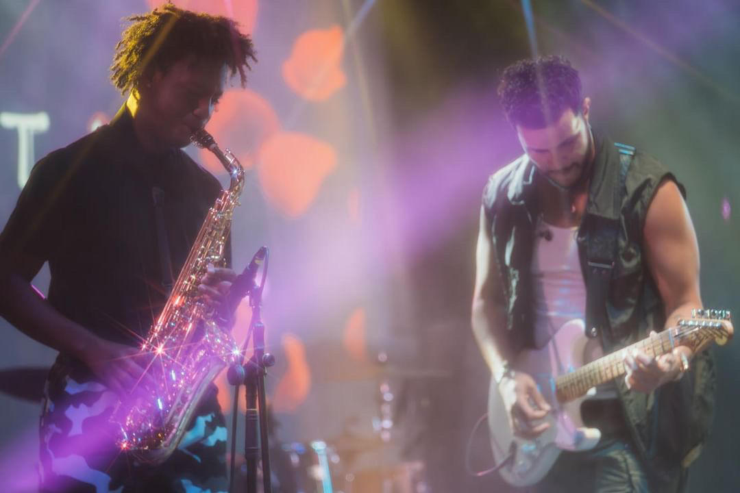 Two musicians perform on stage, one playing saxophone and the other guitar, with colorful stage lighting effects.