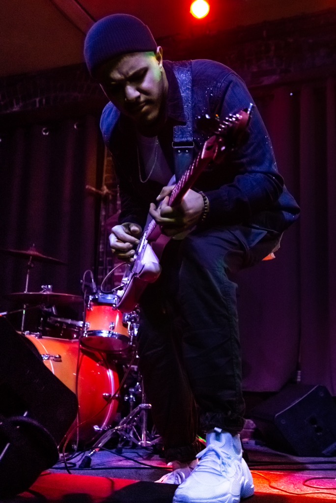 Person playing electric guitar on stage with drum set, dark background, red lighting, wearing beanie and casual clothing.