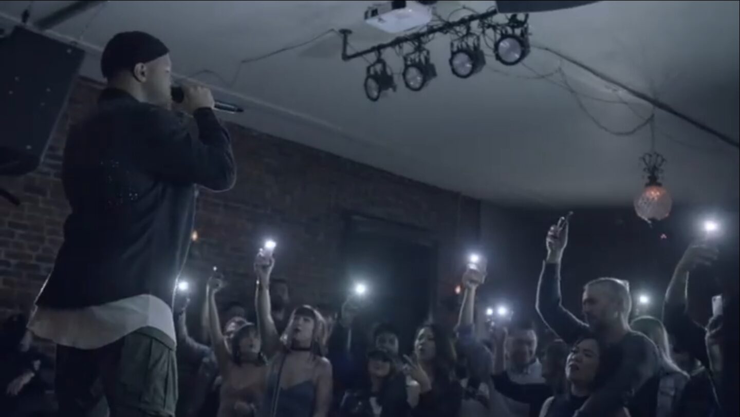 Performer on stage with audience holding up phones in a dark indoor venue.