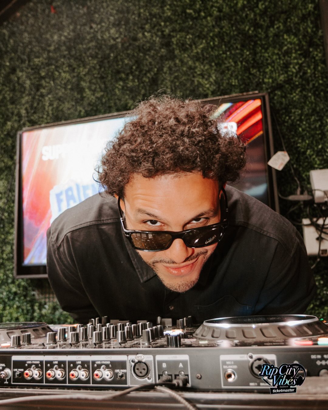 Person with curly hair wearing sunglasses, leaning over DJ equipment, with a screen and green wall in background.