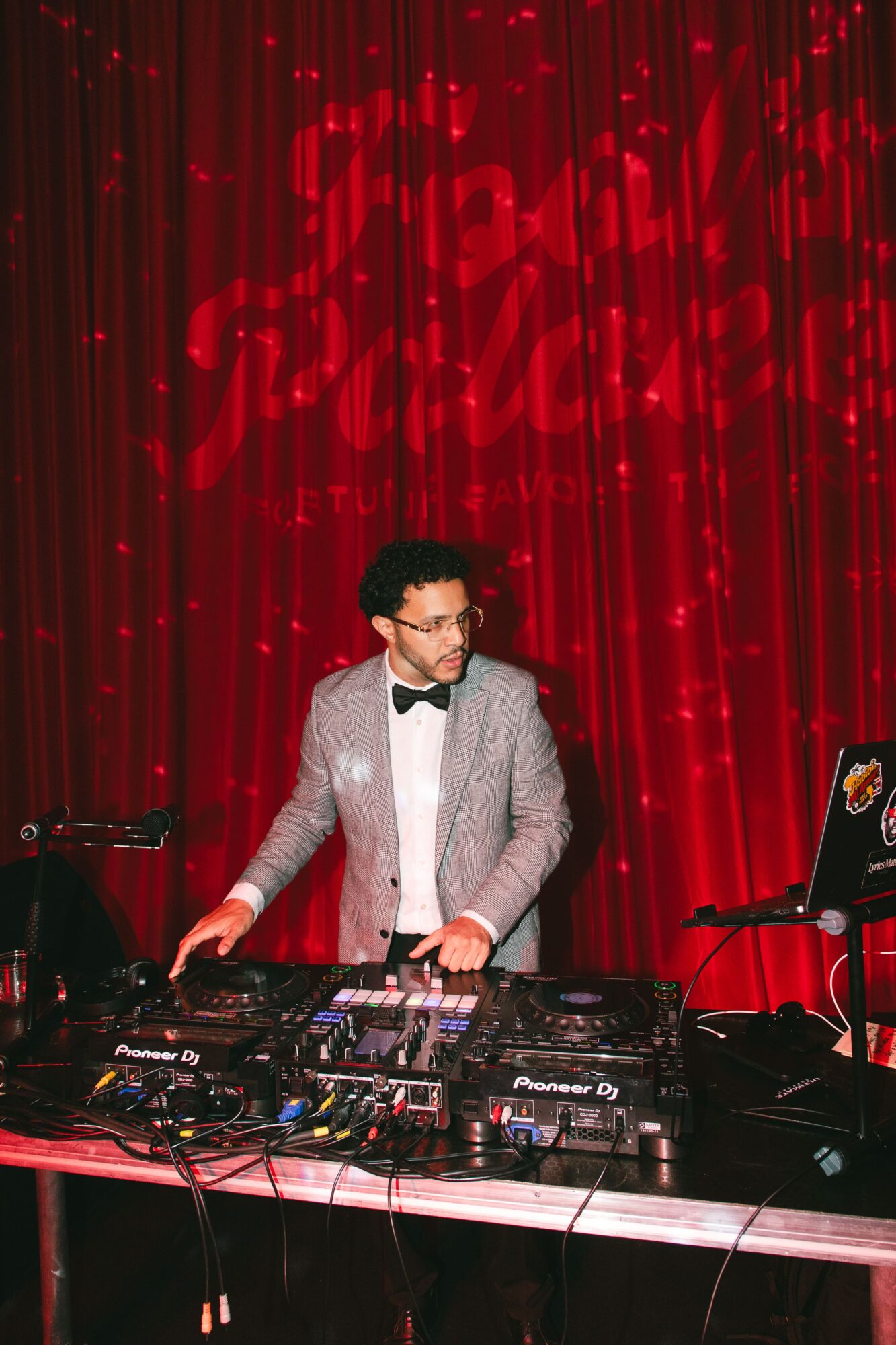 Person in a gray suit and bow tie DJing at a table with equipment, red curtain background, and heart-shaped lights.