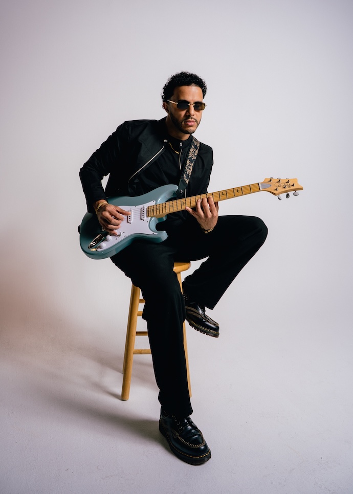 Man sitting on a stool playing an electric guitar, wearing sunglasses and a black jacket, against a plain background.