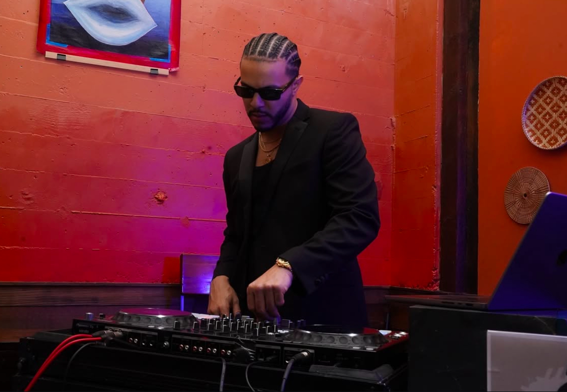 Person with braided hair and sunglasses DJing at a booth in a dimly lit room with colorful wall art.