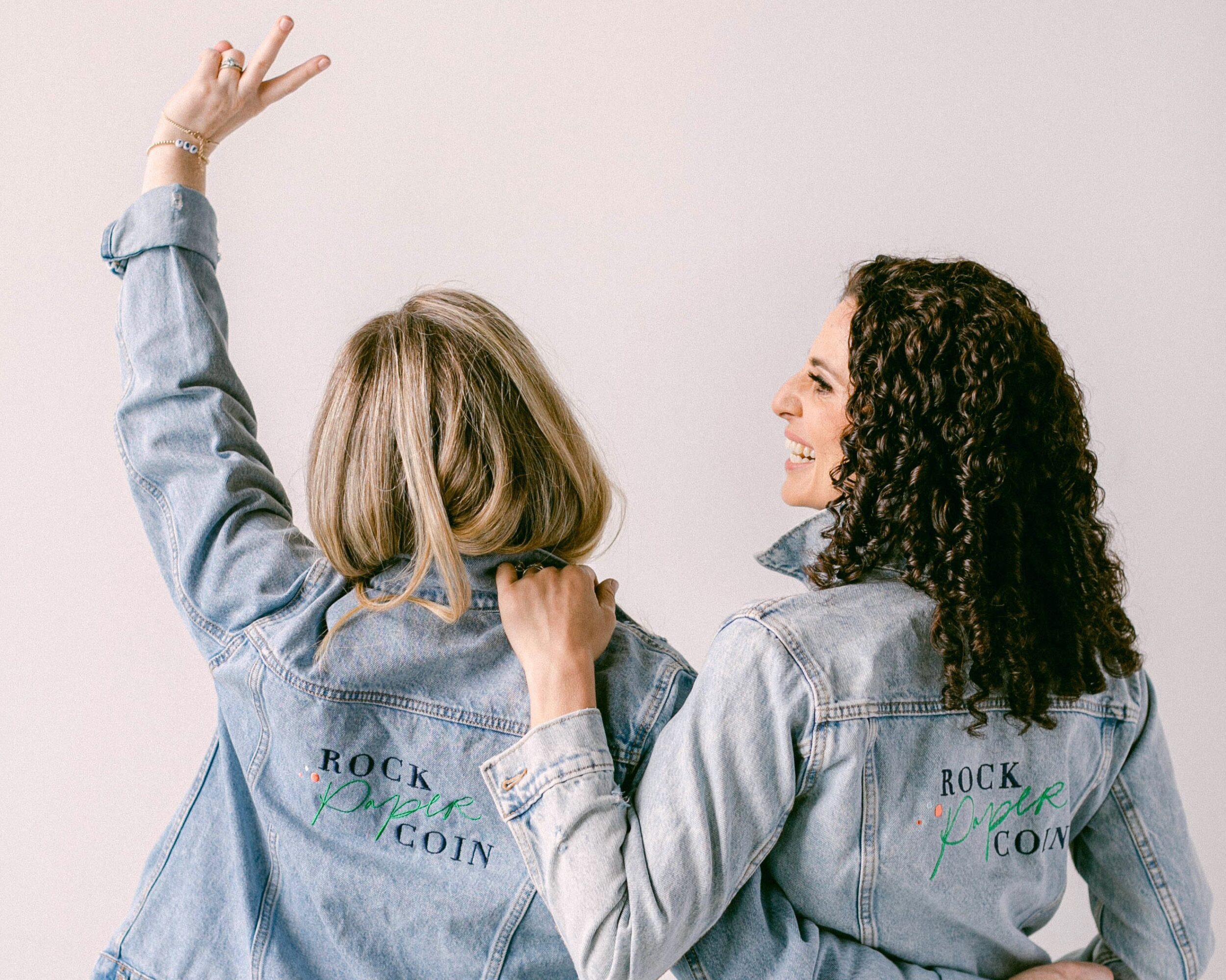 Two women with curly and straight hair wearing denim jackets, smiling and making peace signs, facing each other.