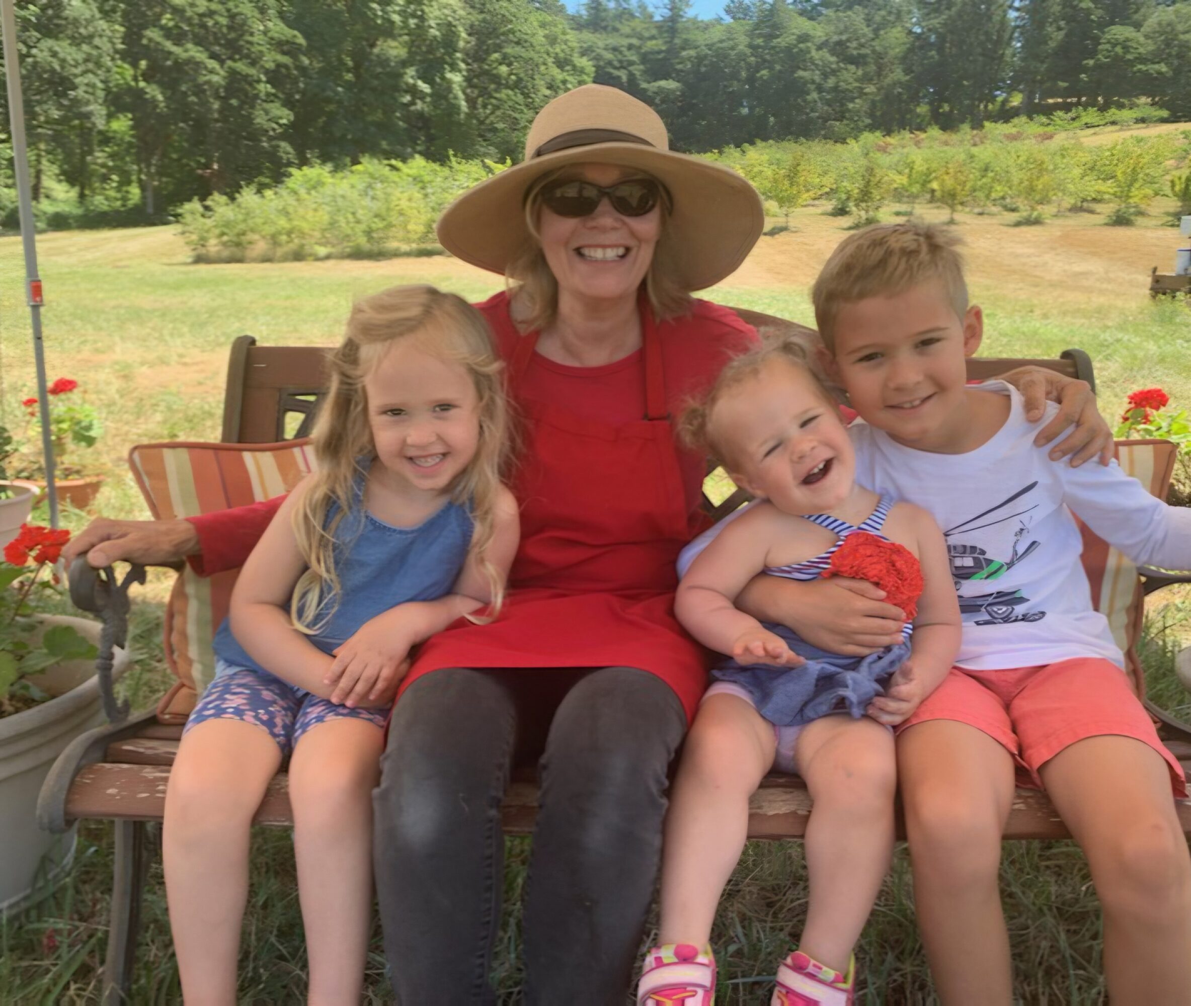 Four people sitting on a bench outdoors, smiling, with trees and a field in the background.