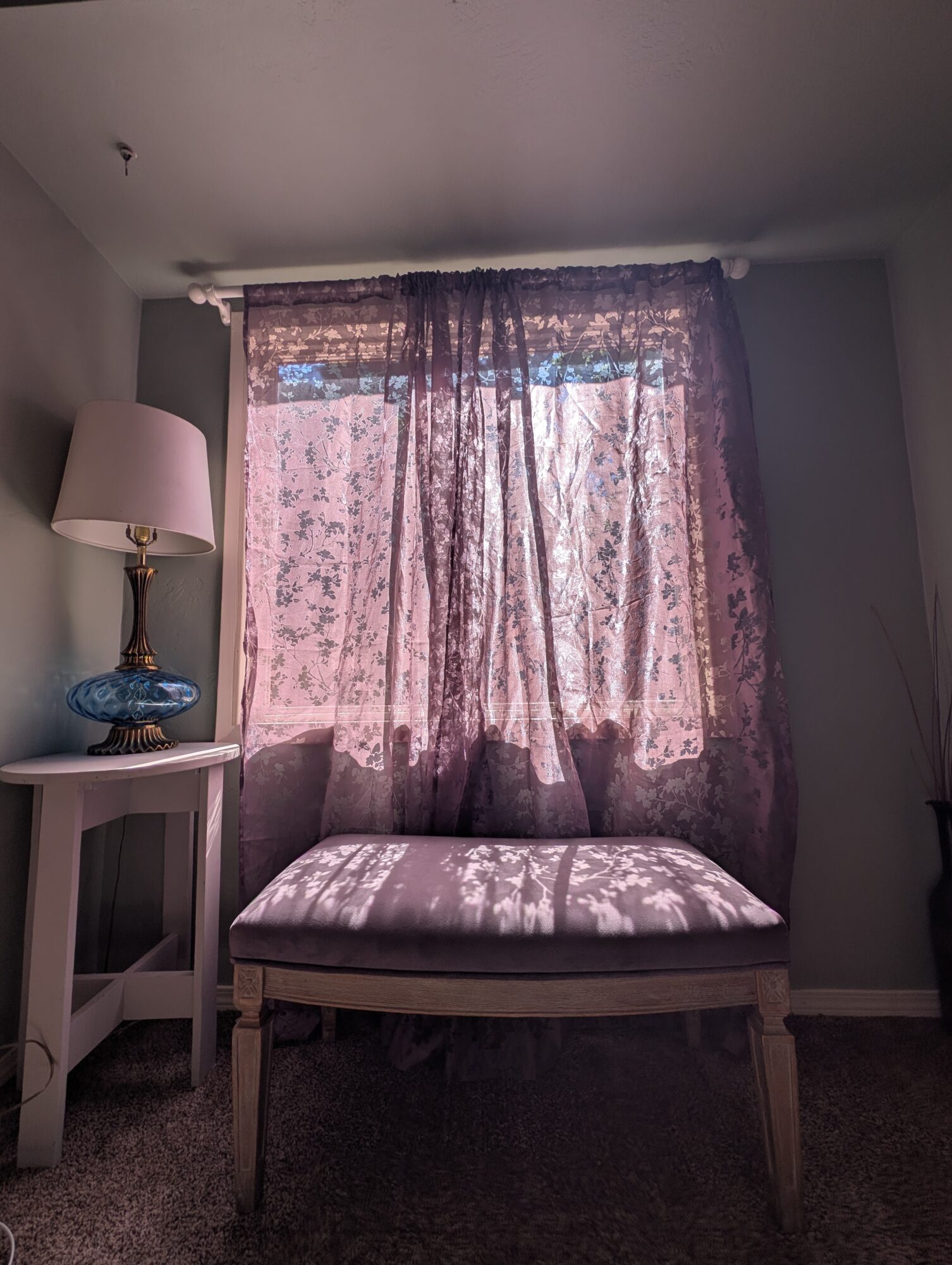Window with pink floral curtains, a cushioned bench, and a side table with a lamp and decorative object.