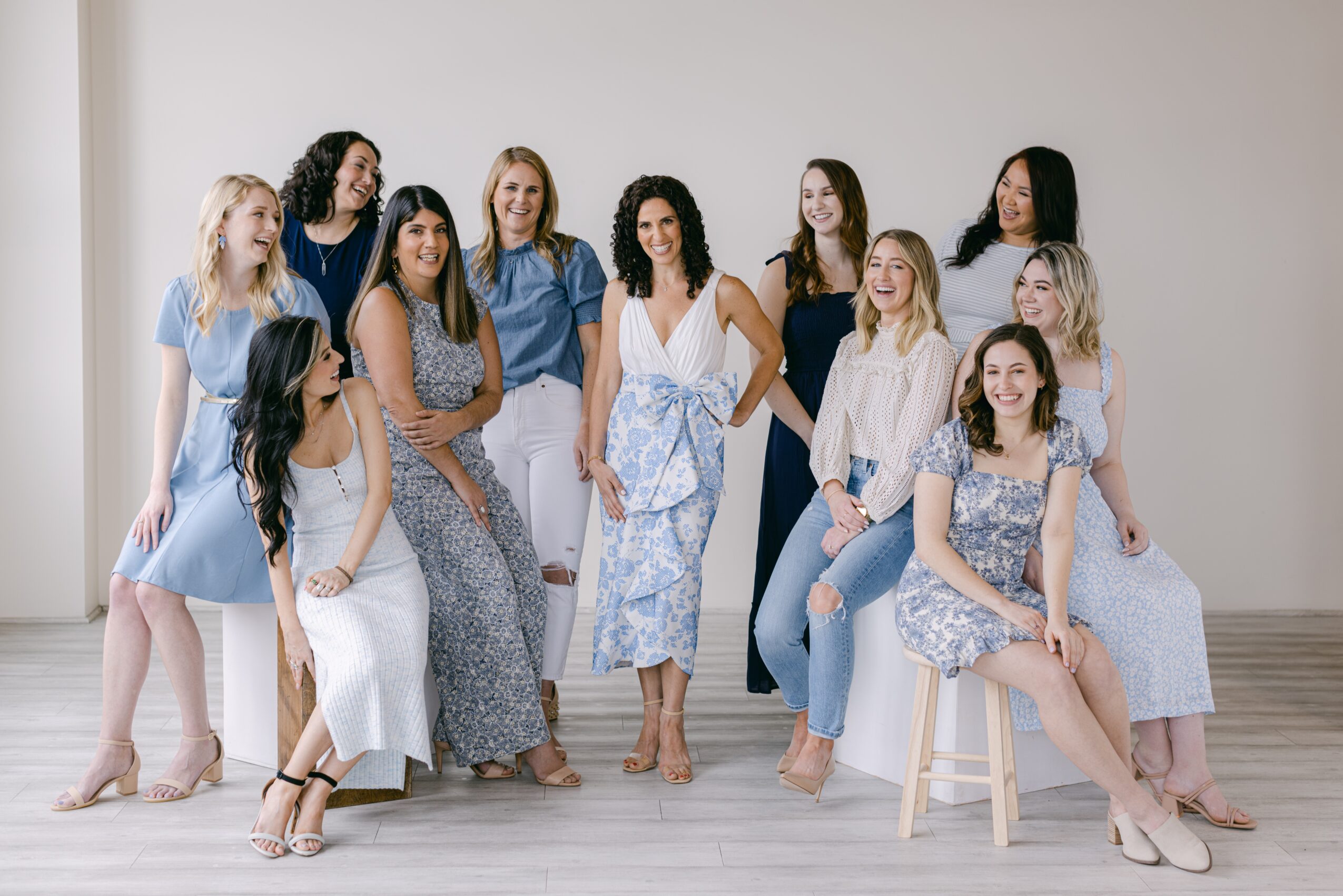 Group of twelve women posing together in a studio, some sitting and some standing, with neutral background.