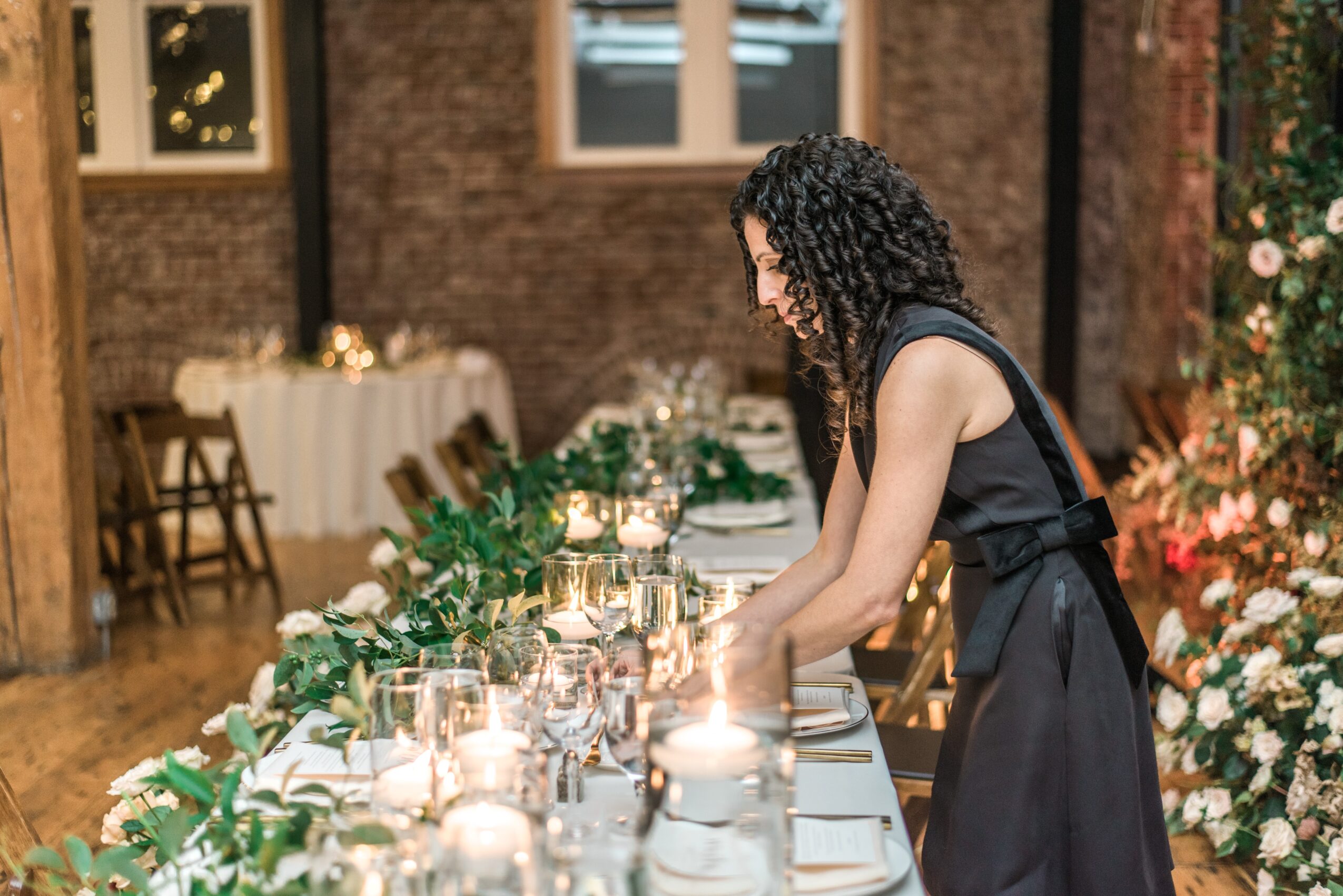 Woman in black dress arranging candles on a decorated table in a warmly lit room with floral arrangements.