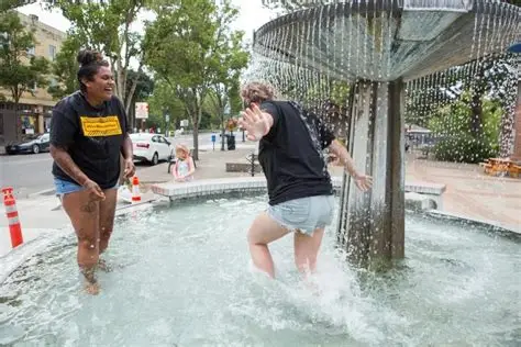 Two people playing in a water fountain, one splashing water, in an outdoor park setting.