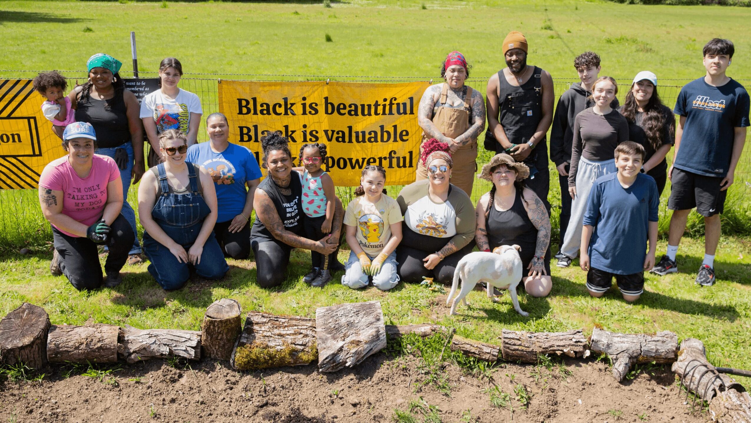Group of people outdoors with a dog, holding a yellow sign, on grassy field with rocks in foreground.