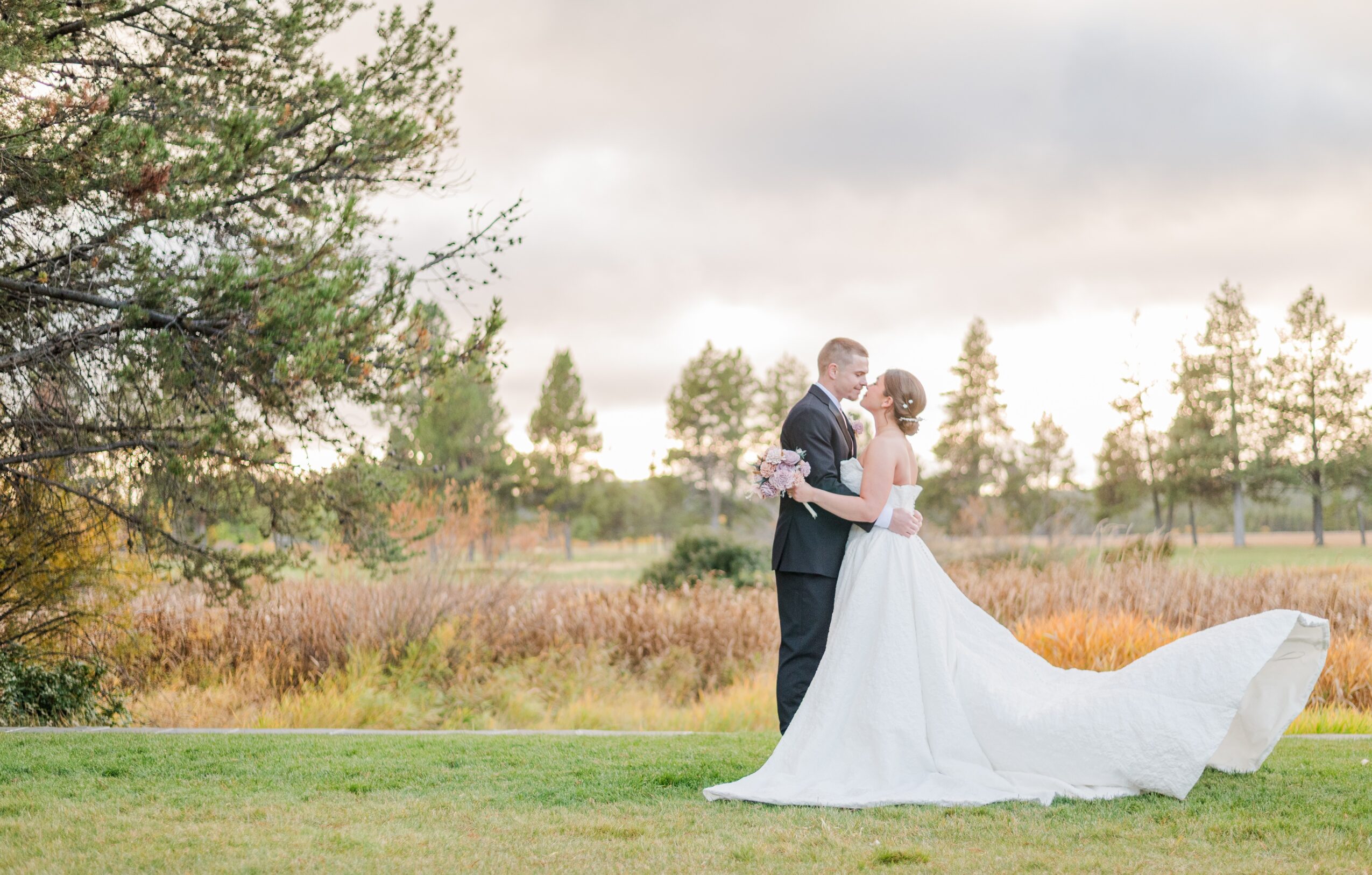 A bride and groom stand close together outdoors, with trees and a cloudy sky in the background.