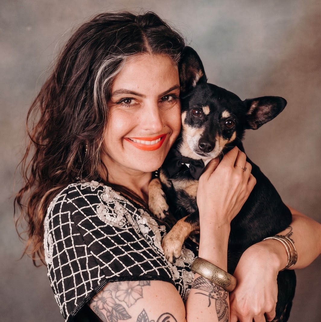 Woman smiling and holding a small black and tan dog, both looking at the camera, woman wearing a patterned top and jewelry.