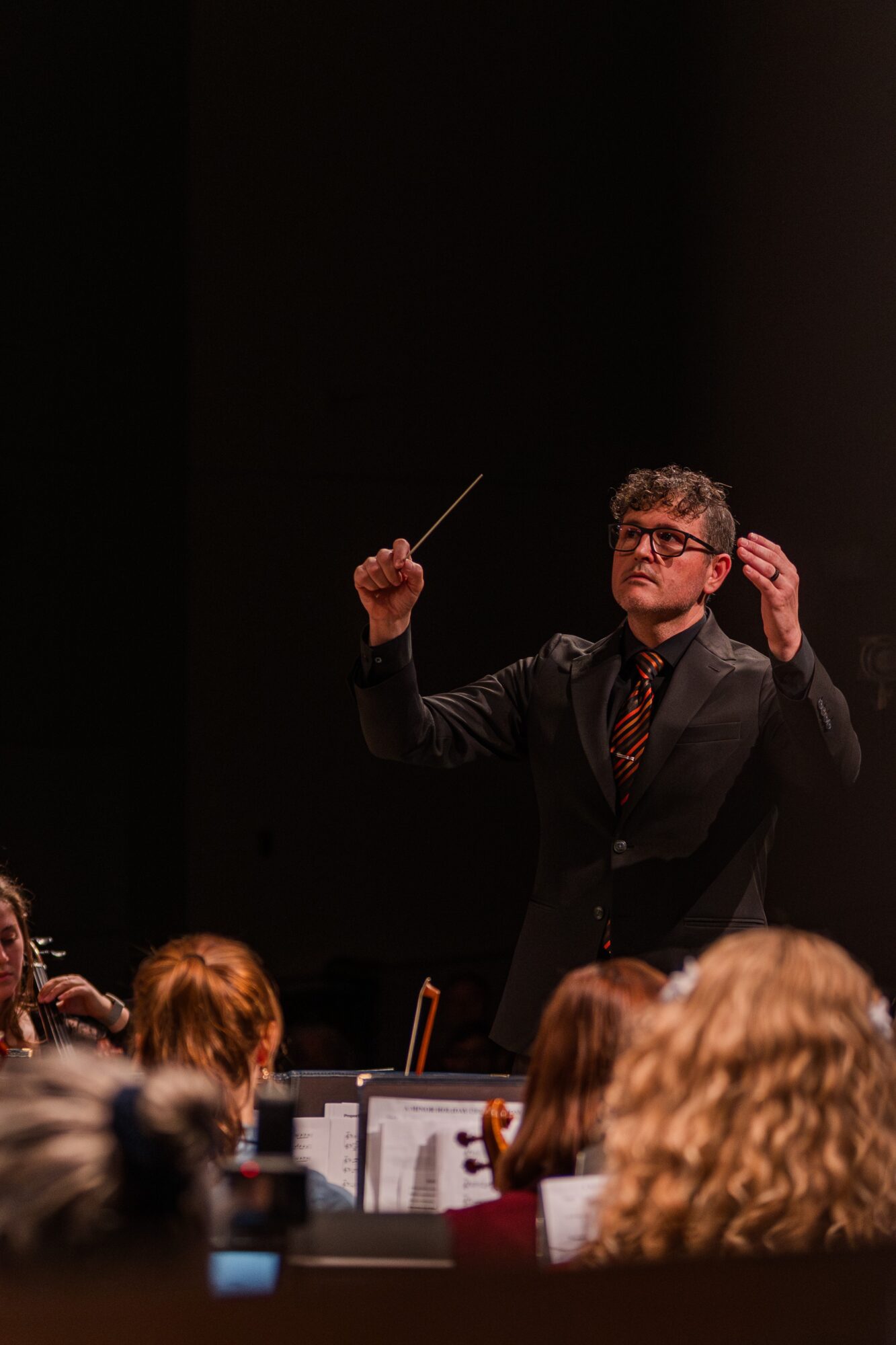 Conductor leading an orchestra on stage with audience in foreground, dark background, conductor wearing glasses and a dark suit, holding a baton.