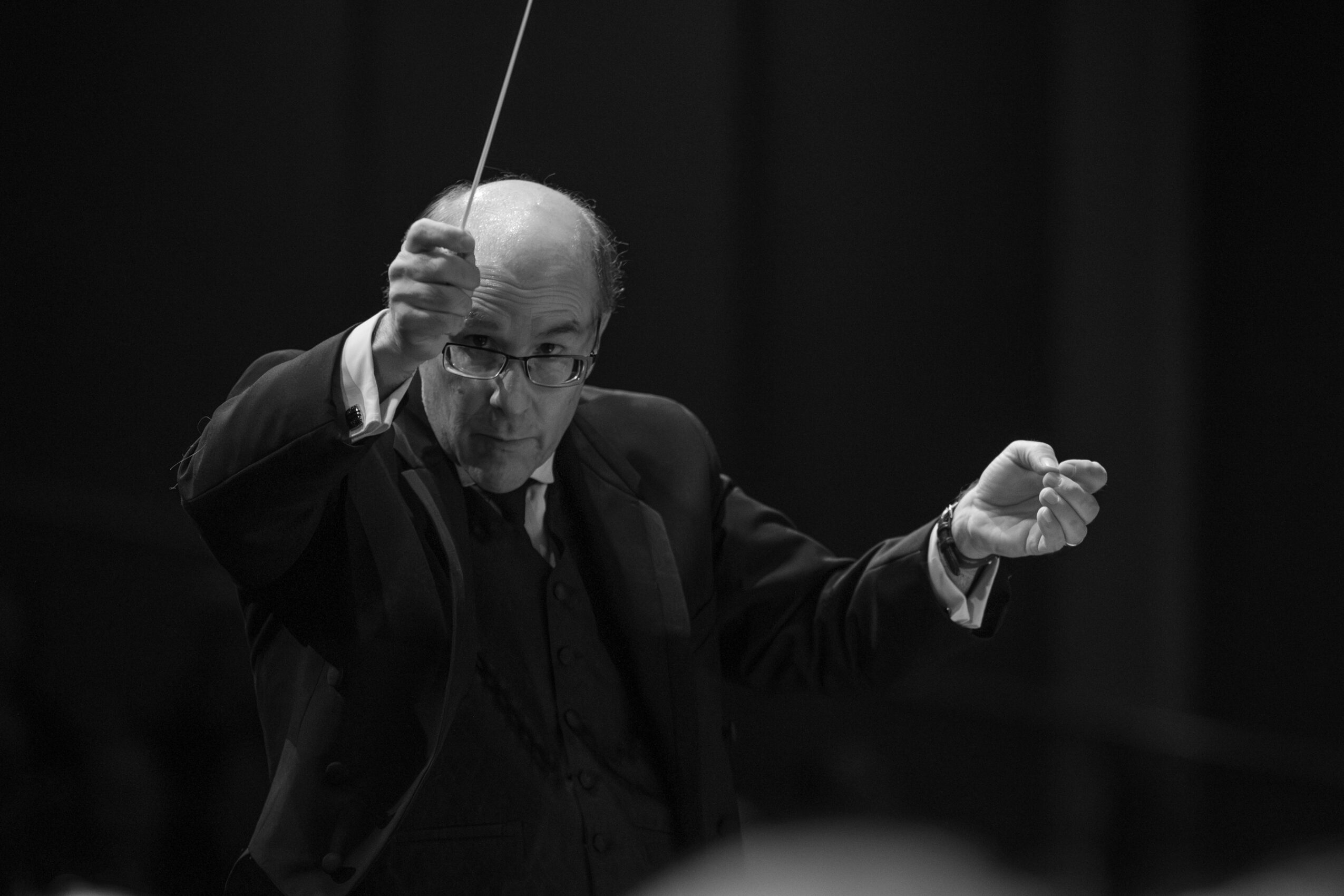 Conductor leading an orchestra, holding a baton, with a focused expression, in a dark setting.