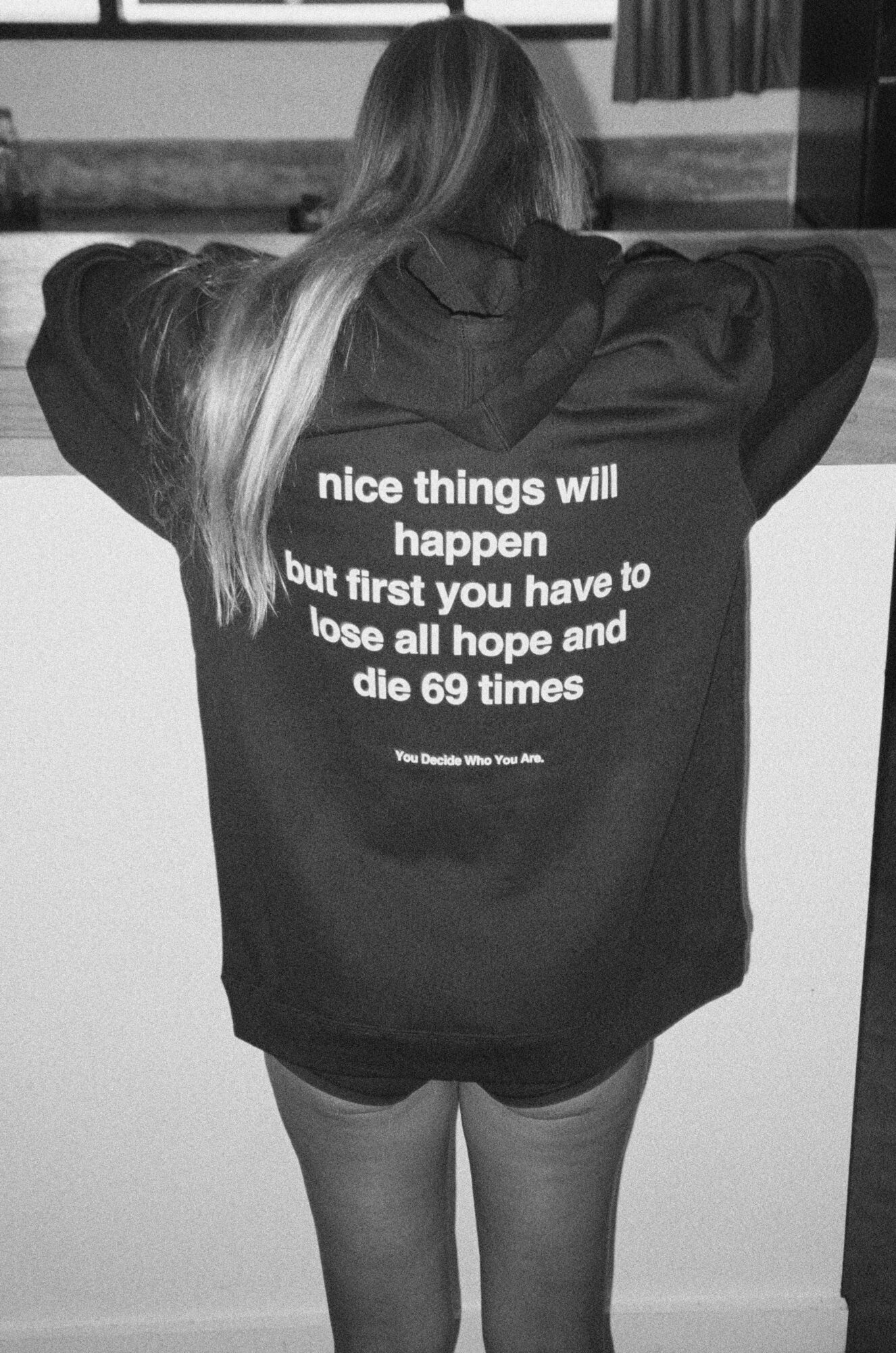 Person with long hair wearing a hoodie with text on the back, standing indoors with arms outstretched.